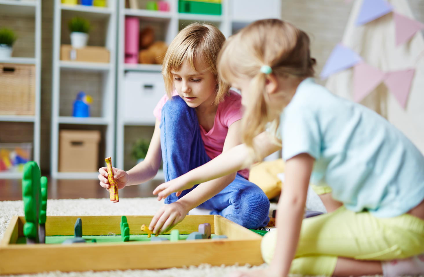 Two young girls playing a board game on the carpet in a playroom with shelves and decorations in the background.