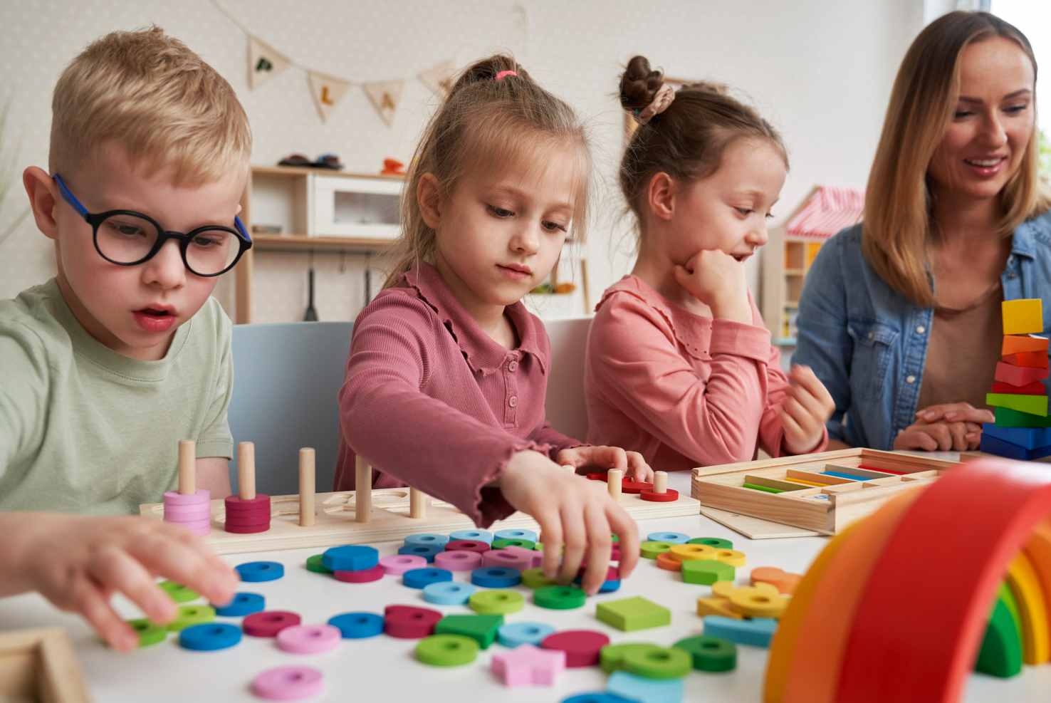 Four children and a woman playing with colorful wooden puzzle pieces and stacking toys at a table in a classroom.