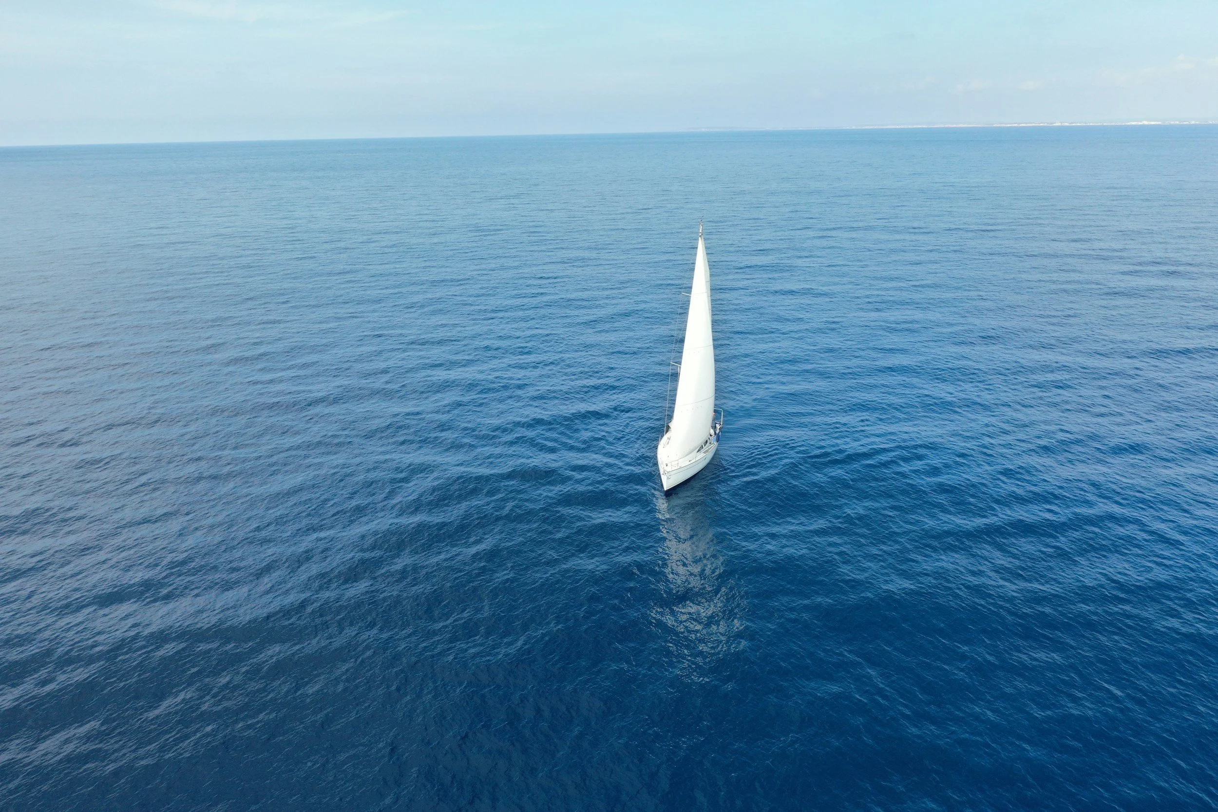 An aerial view of a white sailboat navigating on the open blue ocean under a clear sky.