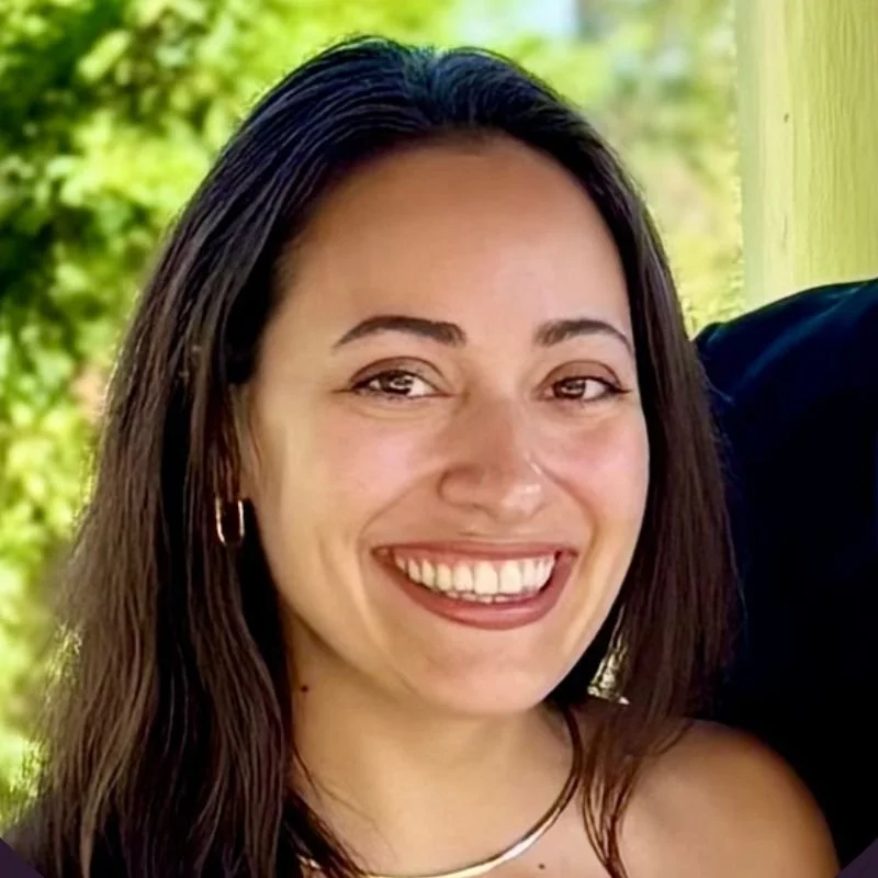 Close-up of a woman smiling outdoors with green foliage in the background.