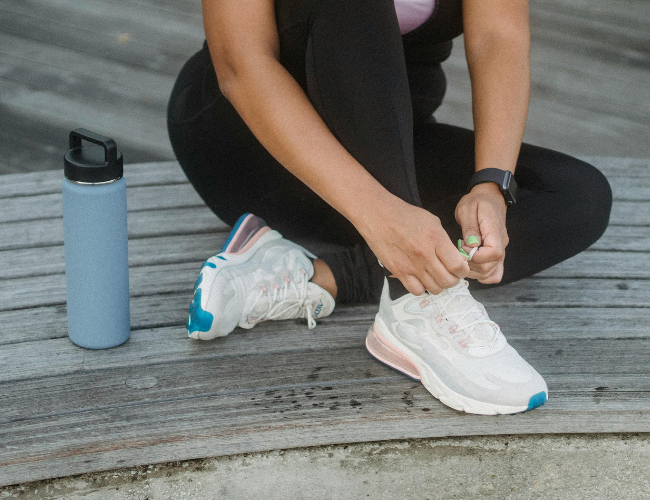 Person sitting on wooden deck tying shoelaces of white athletic shoes with blue and pink accents, next to a blue water bottle.