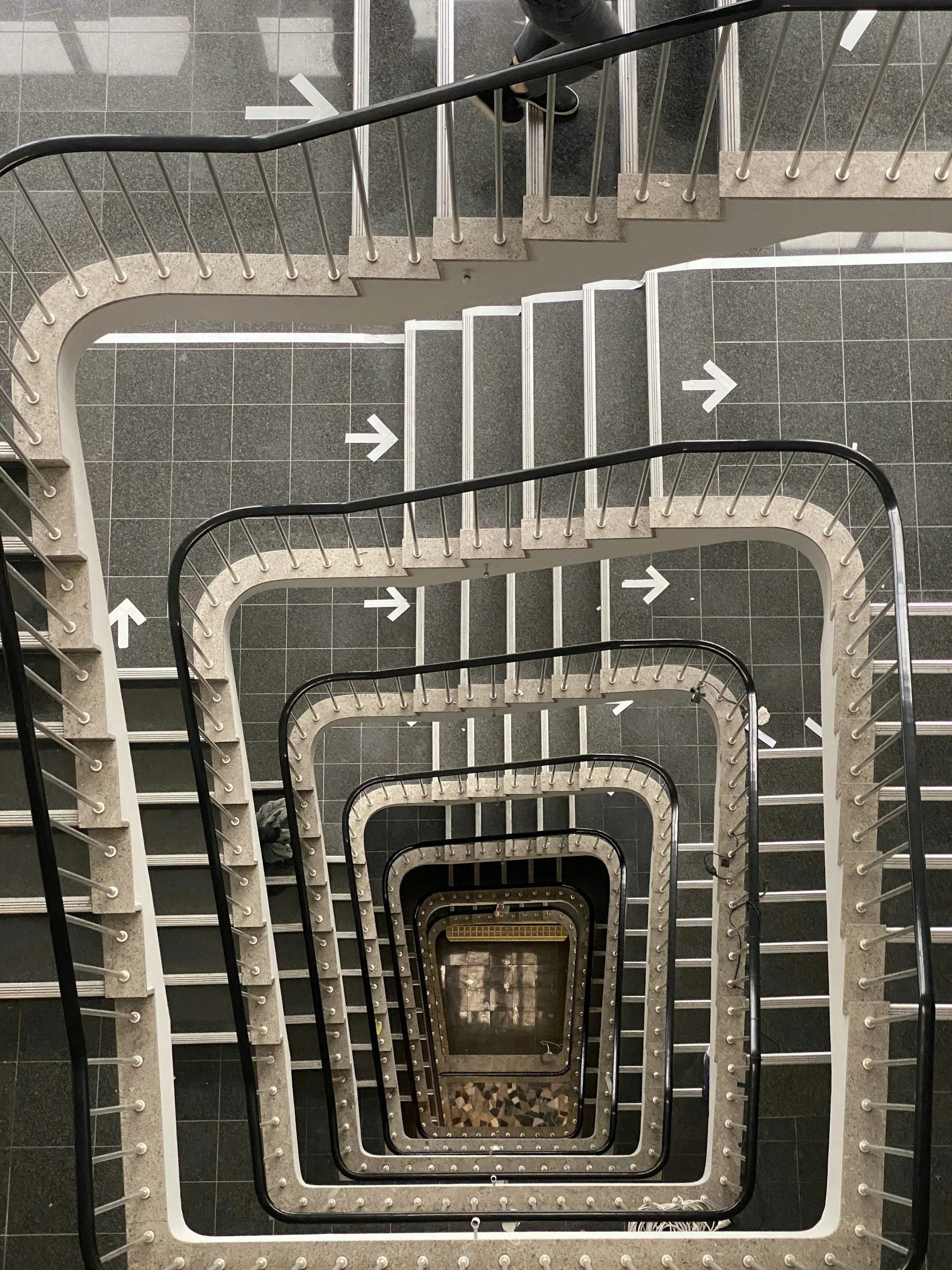 Overhead view of a multilevel staircase with black railings and white steps, spiraling downward with white arrows indicating the direction.