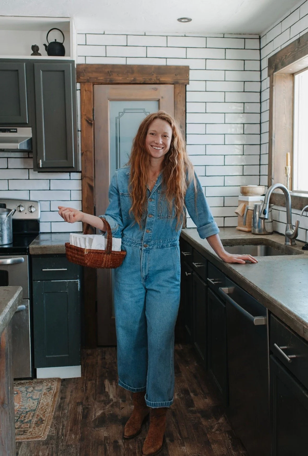 A woman with long red hair wearing a denim jumpsuit and brown boots holding a small basket, standing in a modern kitchen with black cabinets, brick walls, and a window.