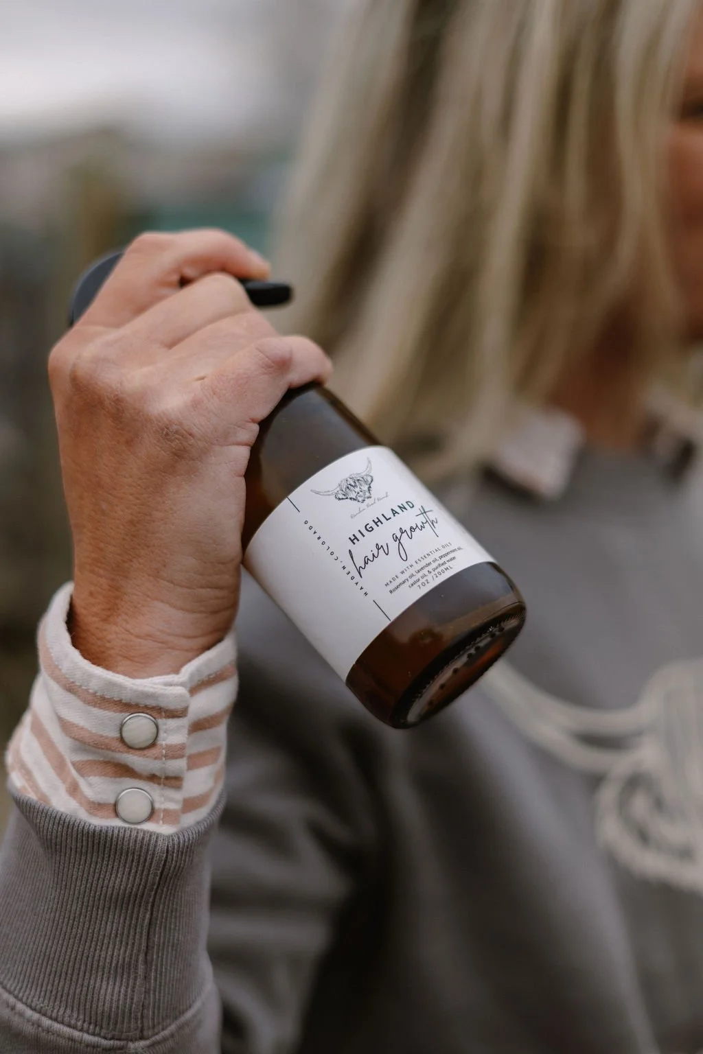 A person holding a brown glass spray bottle labeled 'Highland Hair Growth' with a skull illustration, wearing a striped shirt with buttons on the sleeve.