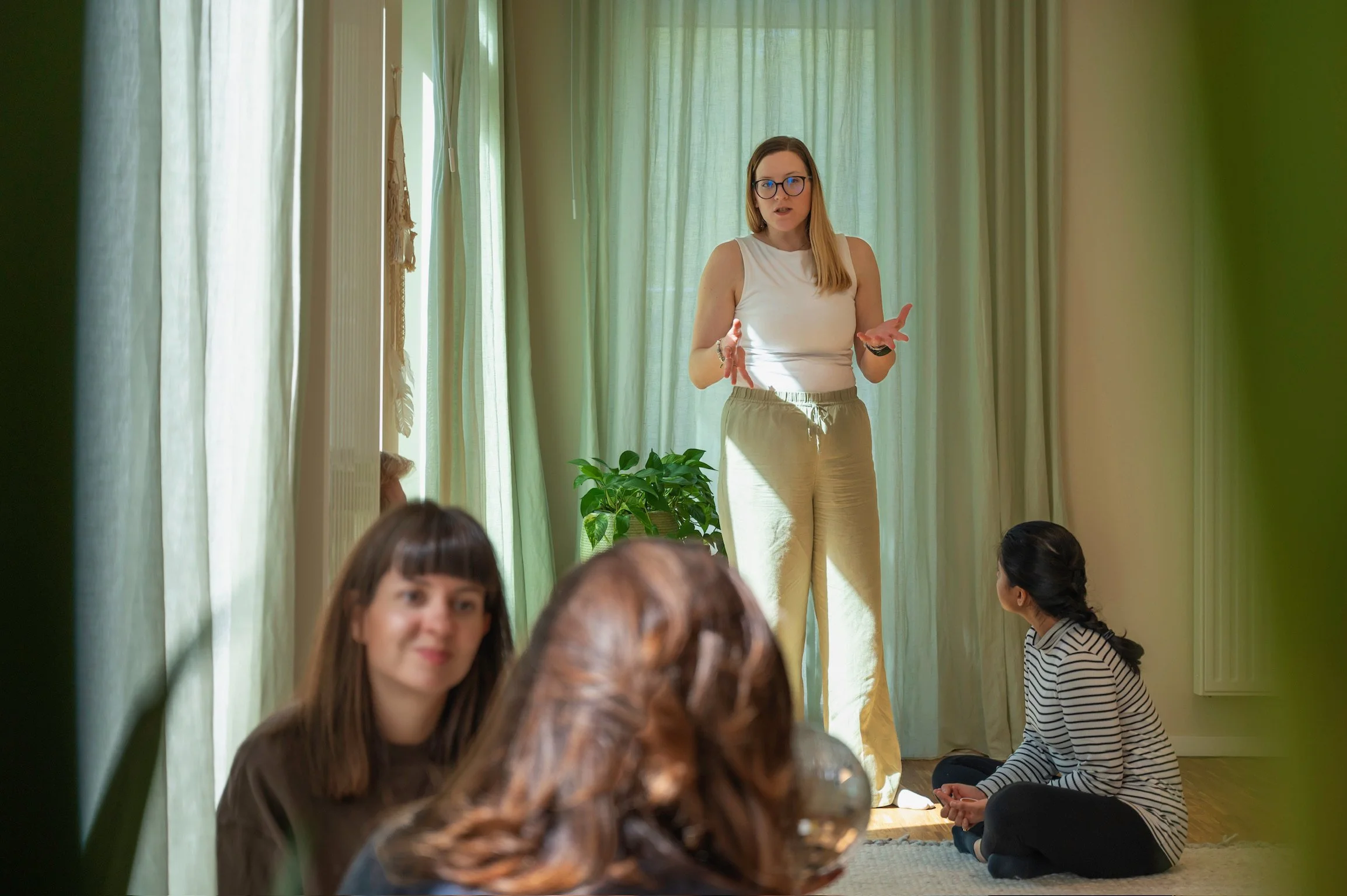A woman in glasses and beige pants is speaking in front of a small group of women sitting on the floor, with green curtains behind her and a potted plant nearby.