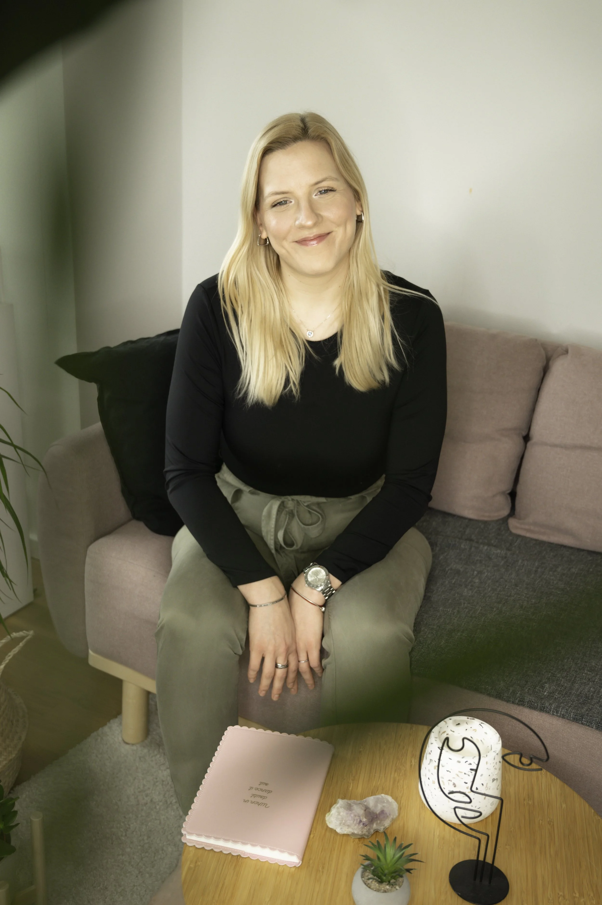 A woman with long blonde hair, wearing a black long-sleeve shirt and khaki pants, sitting on a pink sofa in a cozy living room, smiling at the camera.