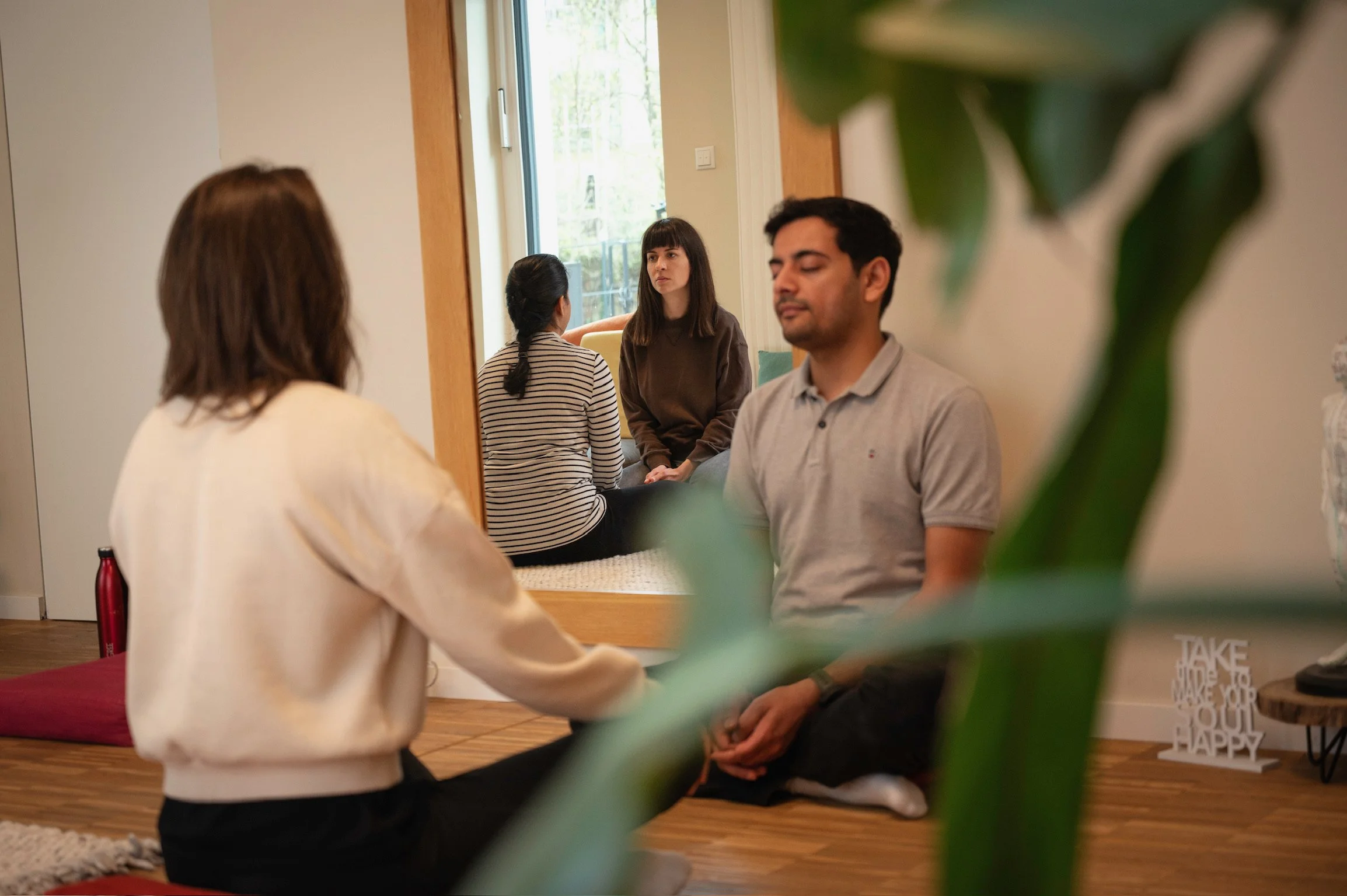 A group of five people in a therapy or meditation session, seated on the floor in a cozy room with natural light, some with eyes closed, others listening attentively.