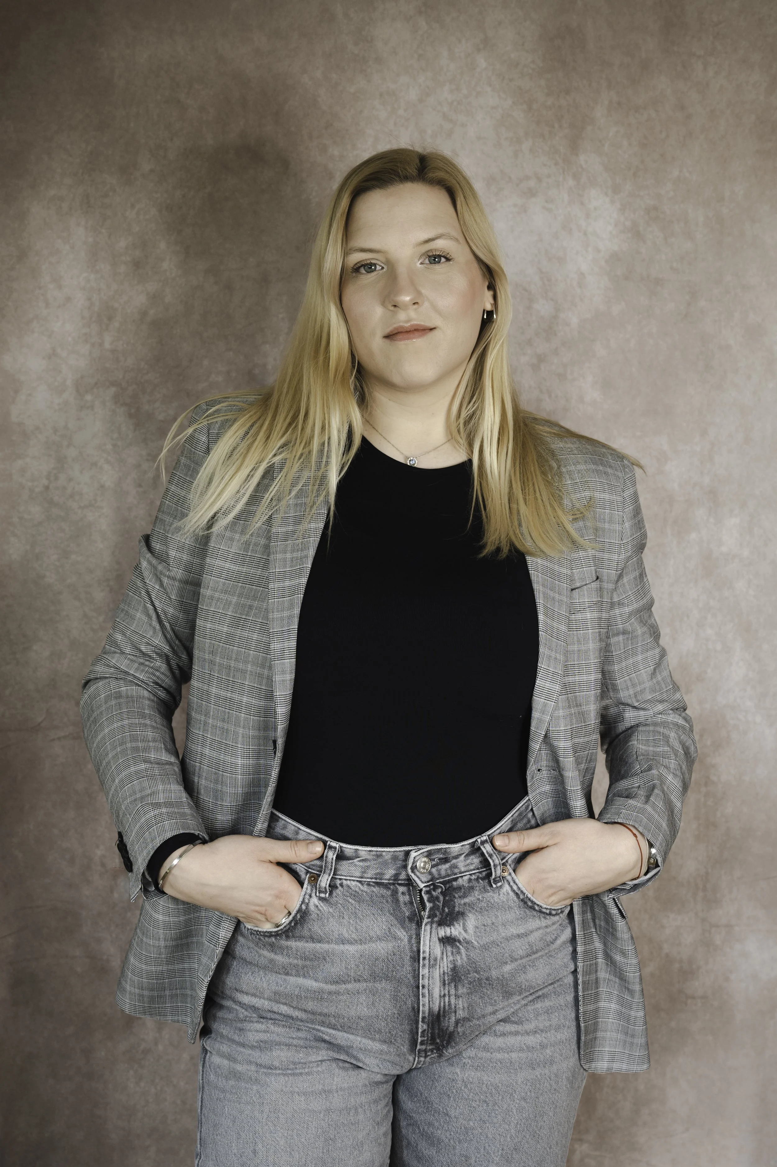 A young woman with blonde hair wearing a gray plaid blazer, black shirt, and gray jeans standing against a textured beige background.