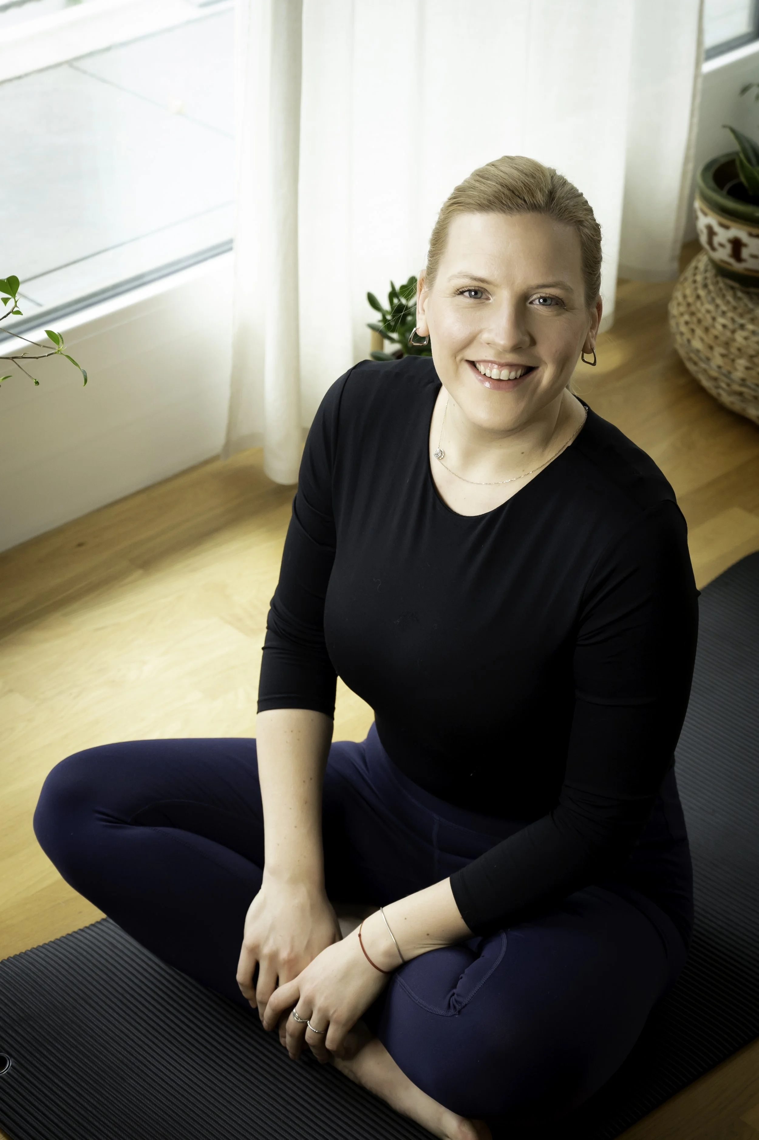 A woman smiling and sitting cross-legged on a yoga mat near a window, wearing a black long-sleeve shirt and navy blue pants.