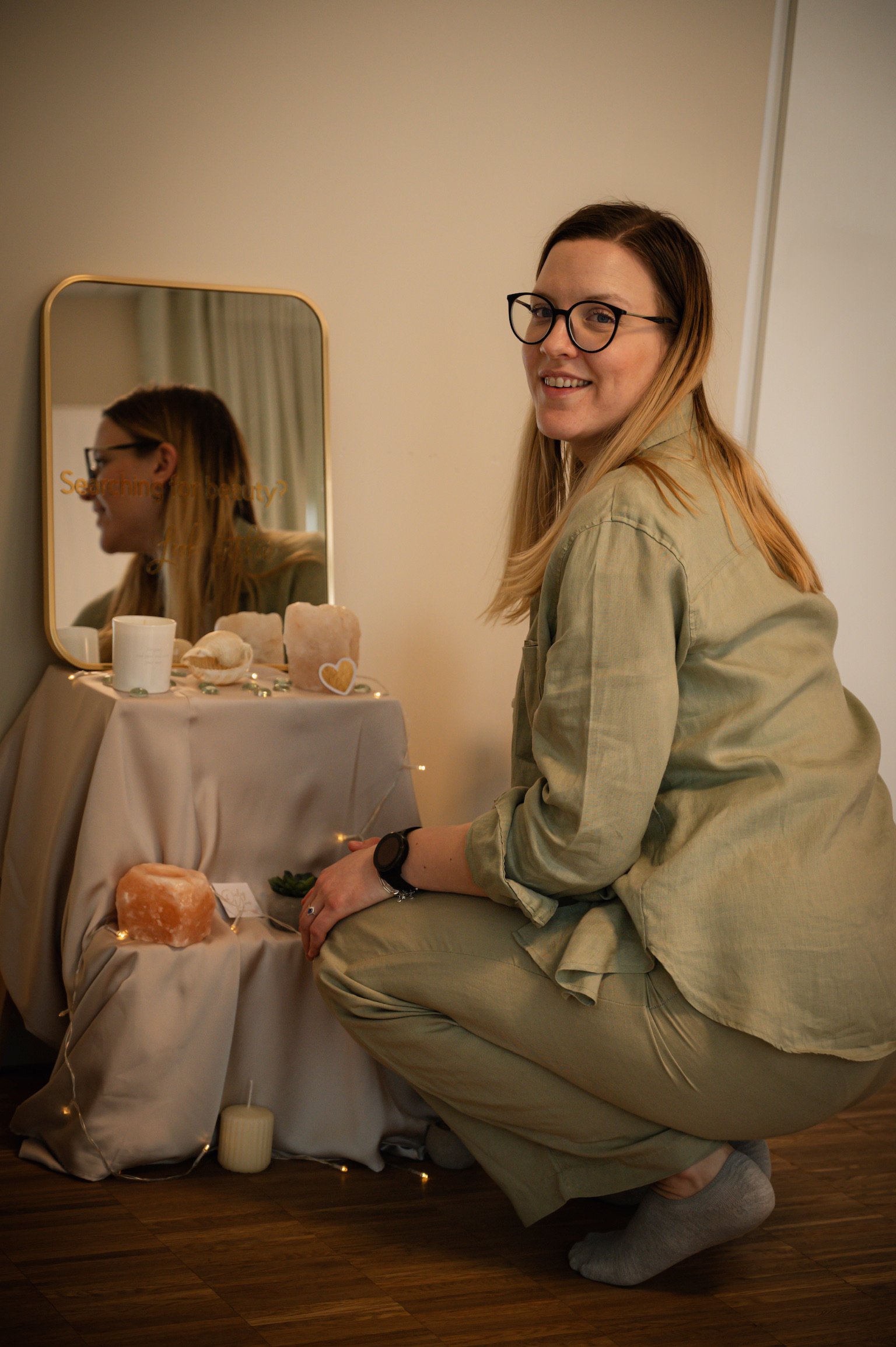 A woman with long blonde hair and glasses kneeling on the floor next to a small table with candles, rocks, and decorative items. She is smiling and looking at the camera.