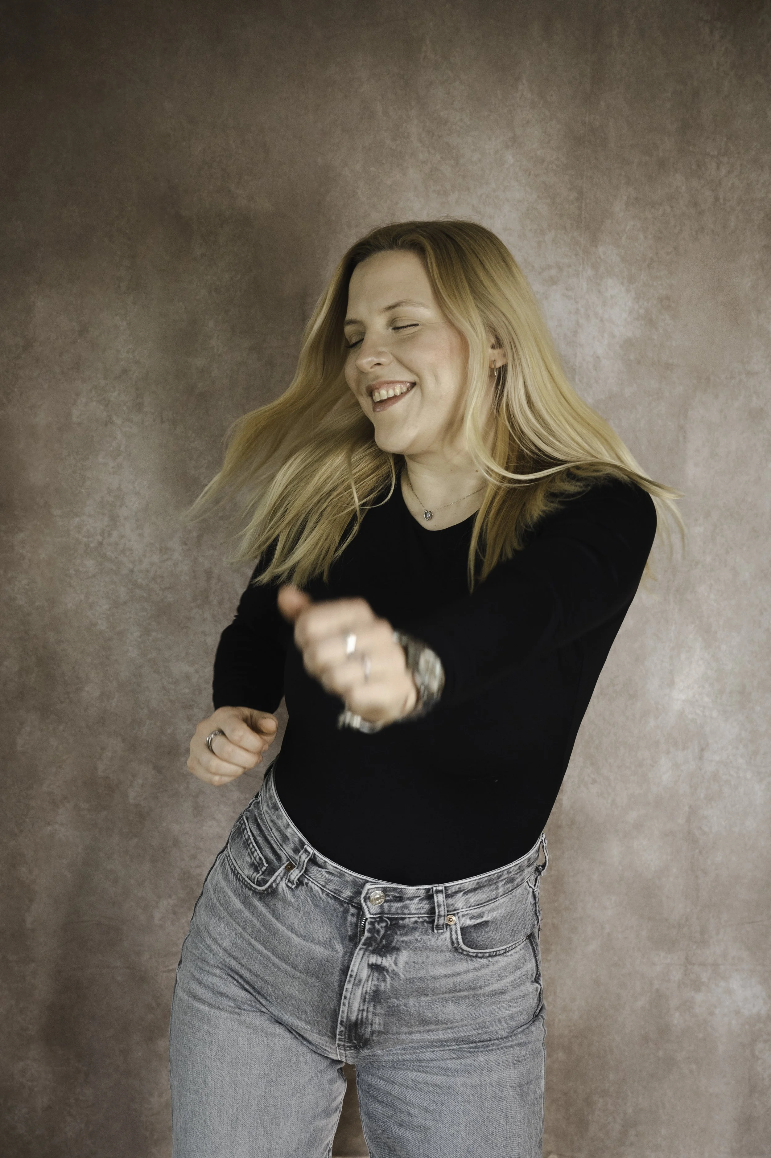 A woman with blonde hair, wearing a black long-sleeve shirt and light gray jeans, smiling and dancing against a textured beige background.