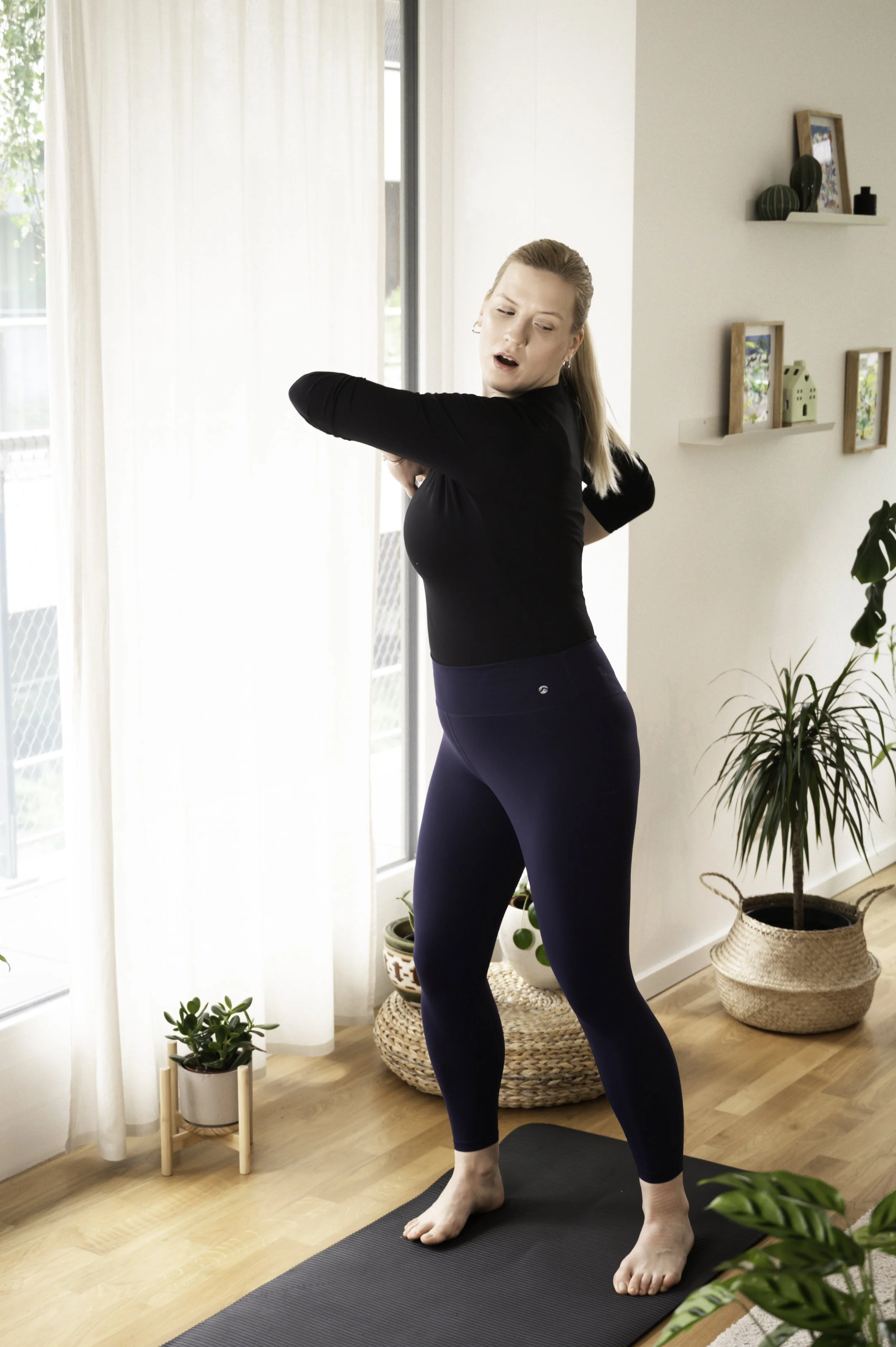 Woman in black shirt and blue leggings stretching indoors on a yoga mat near large window with white curtains, surrounded by potted plants and wall shelves.