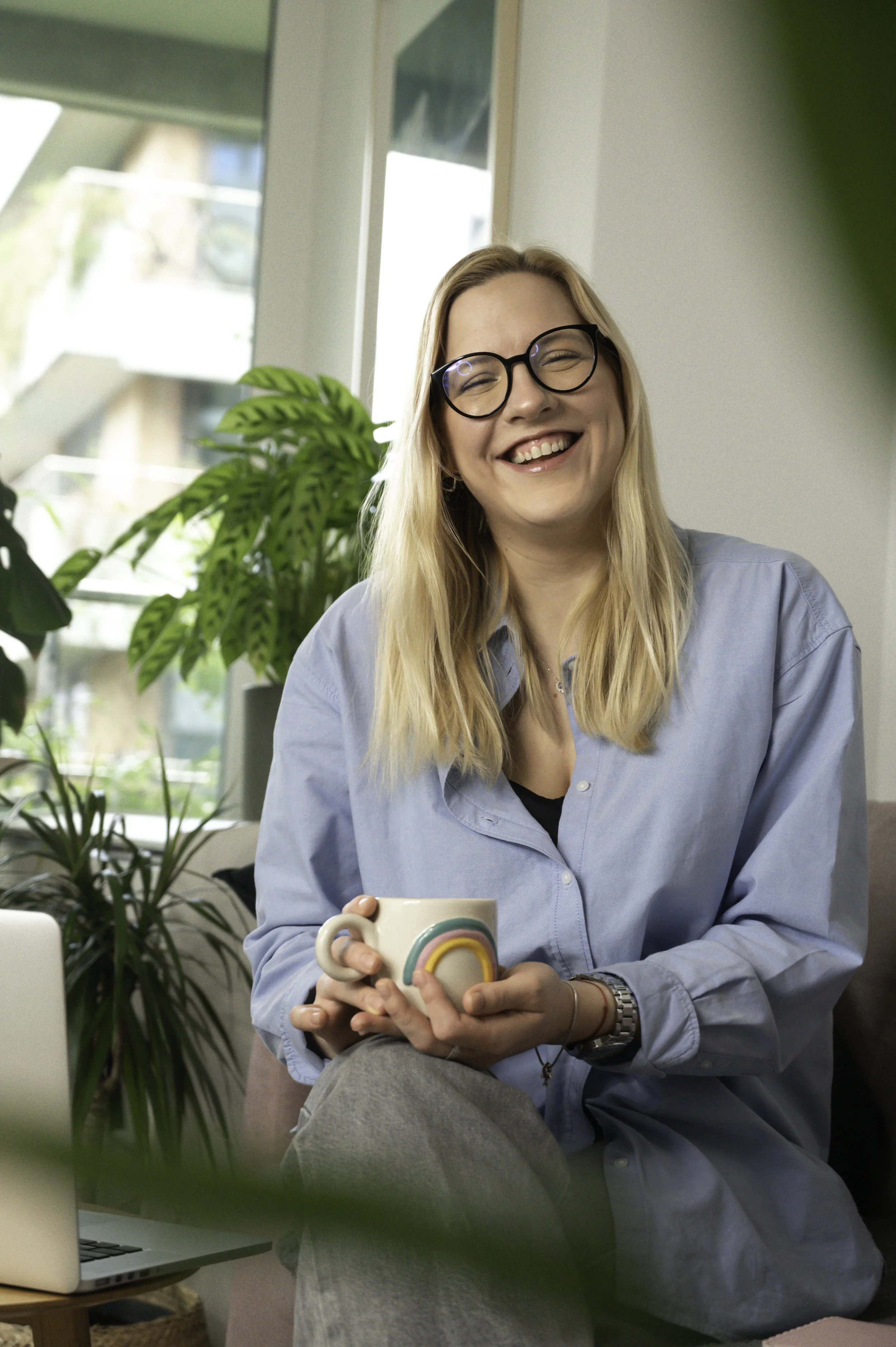 A woman with blonde hair, glasses, and a blue shirt smiling and sitting on a couch in a bright room with plants, holding a mug with a rainbow design.