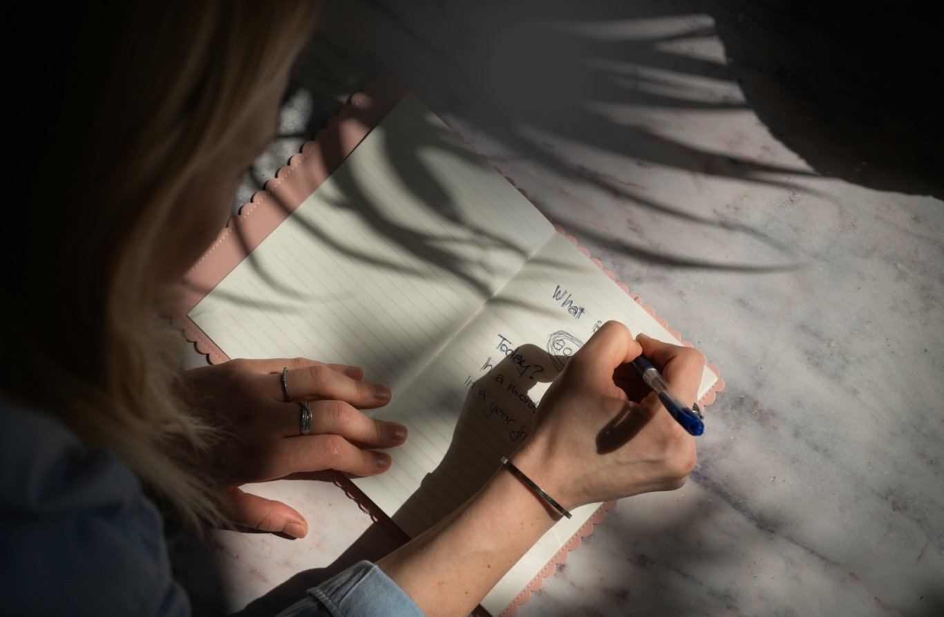 A person writing in a notebook with lined pages, sitting at a marble table. The person is holding a blue pen and is in focus, with sunlight casting shadows on the table.