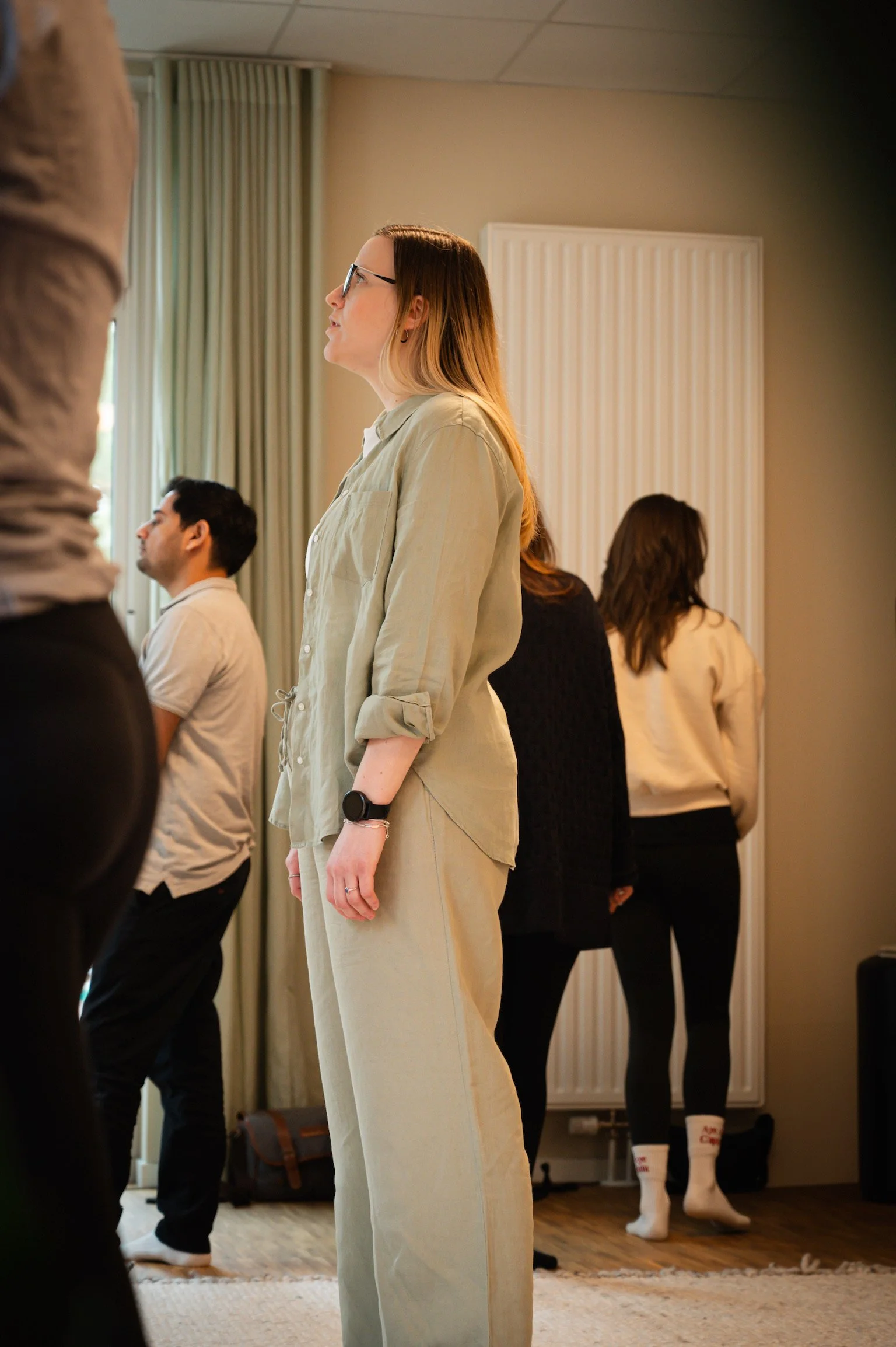 A group of people standing in a room, with a woman wearing glasses and beige clothing in the foreground, and others behind her.