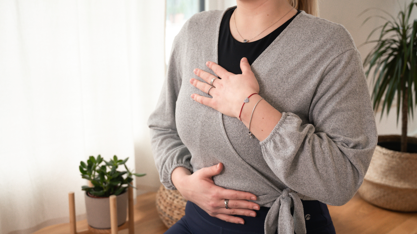 A woman showing a gesture of having her hand on her chest and stomach, possibly indicating breath or emotion, in a cozy home setting with plants and natural light.