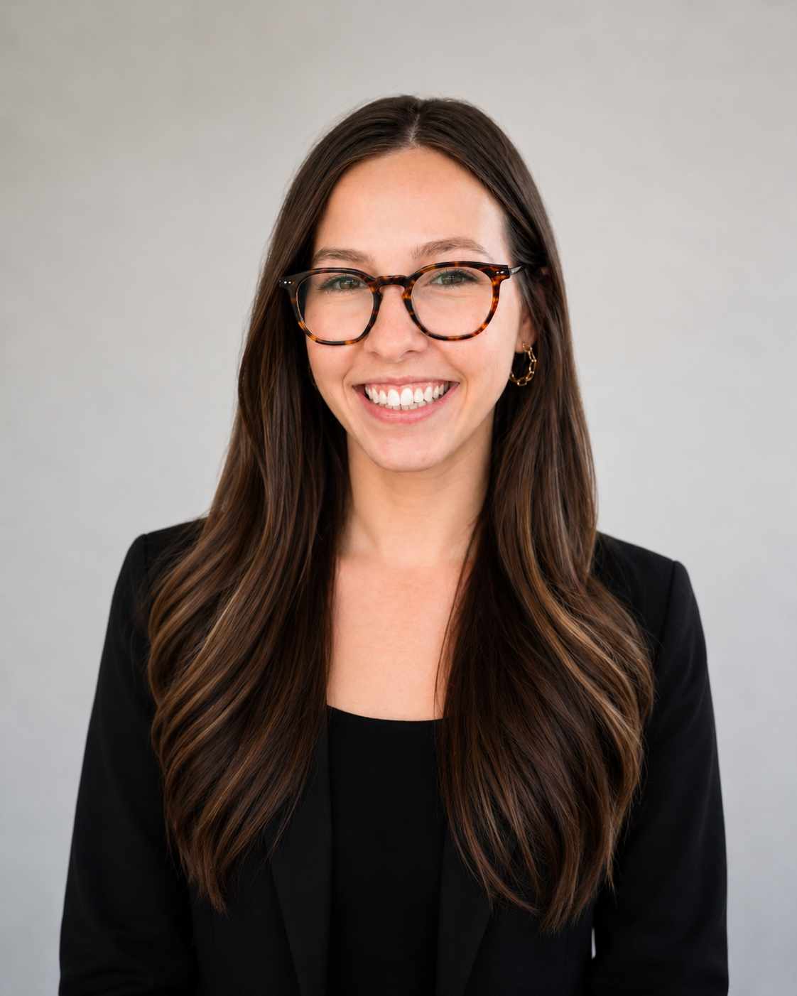 A smiling woman with long wavy brown hair, wearing glasses, a black blazer, and a red shirt, standing against a plain white background.