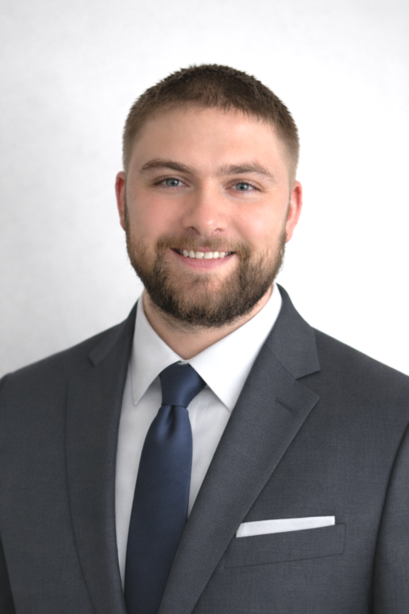 Portrait of a smiling man with a beard and short hair, wearing a gray suit, white shirt, and dark blue tie, against a plain light background.
