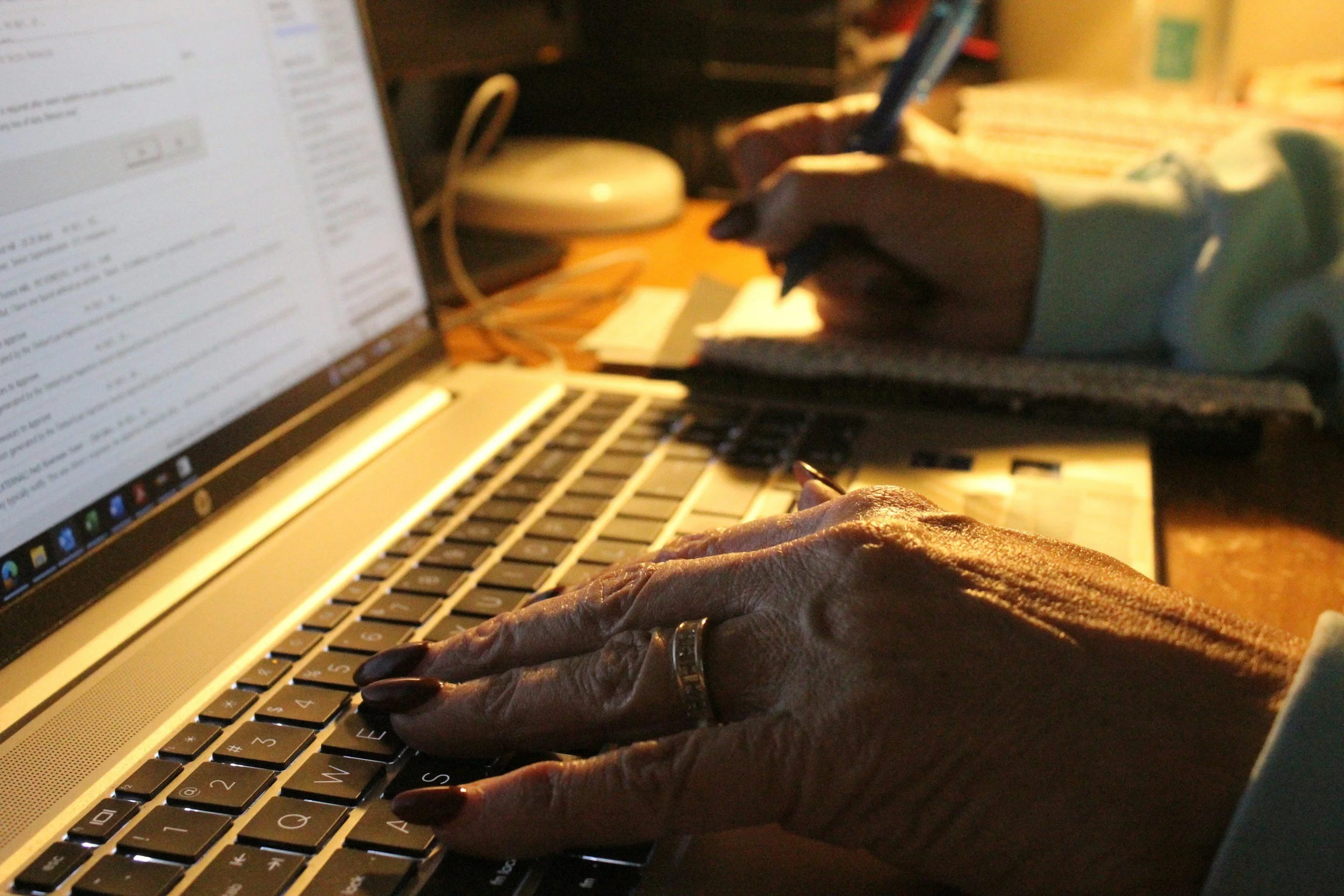 Close-up of elderly woman’s hands typing on a laptop keyboard with a ring on her finger and a pen in her hand, on a cluttered desk with papers, a notebook, and a lamp.