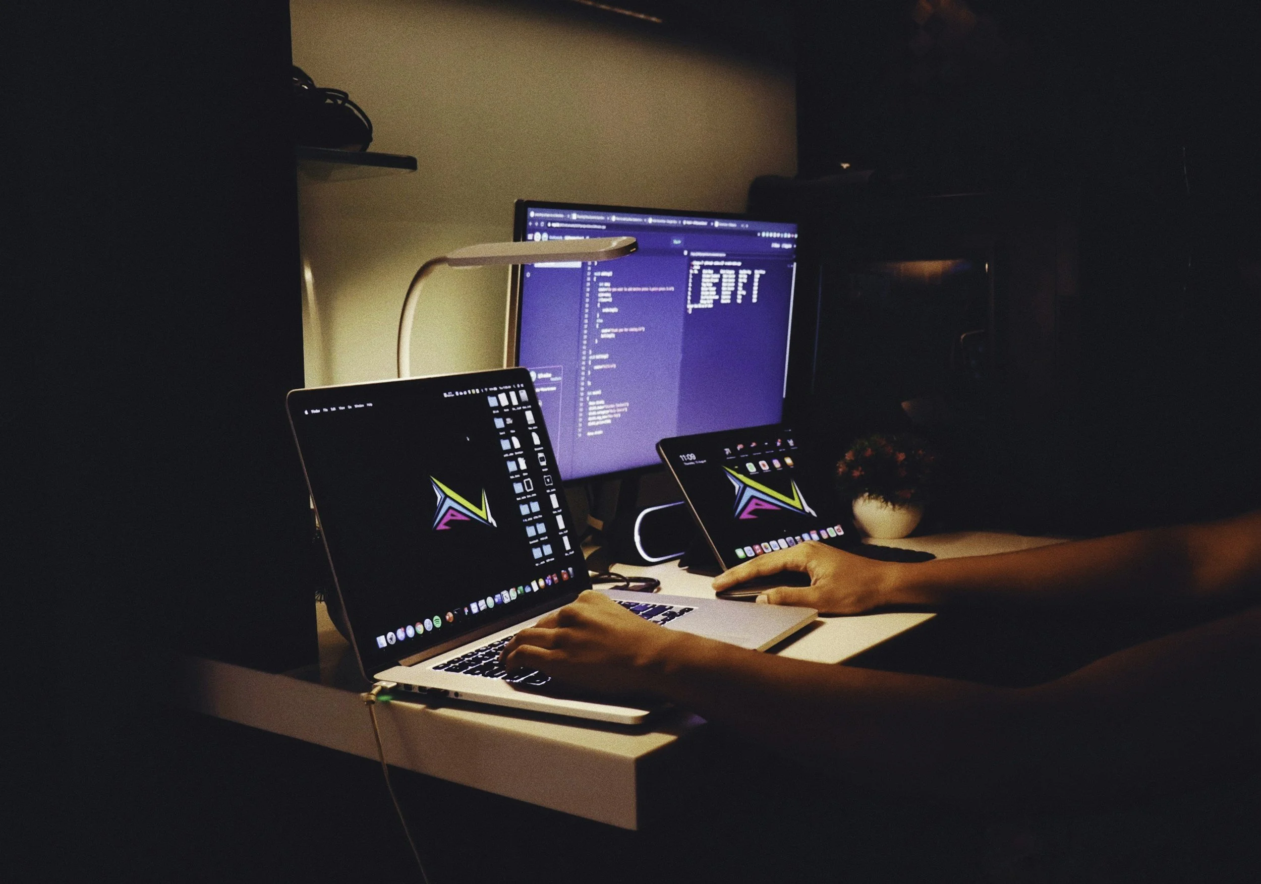 A workspace with a person using two laptops and a desktop monitor, all displaying colorful graphics and coding interfaces, illuminated by a white desk lamp in a dimly lit room.