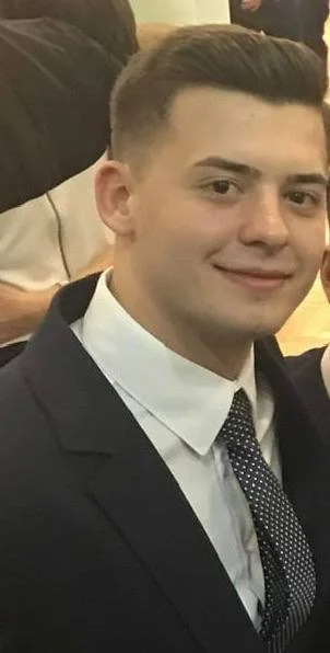 Young man in formal attire smiling at the camera, wearing a suit, white shirt, and tie, at a social event.