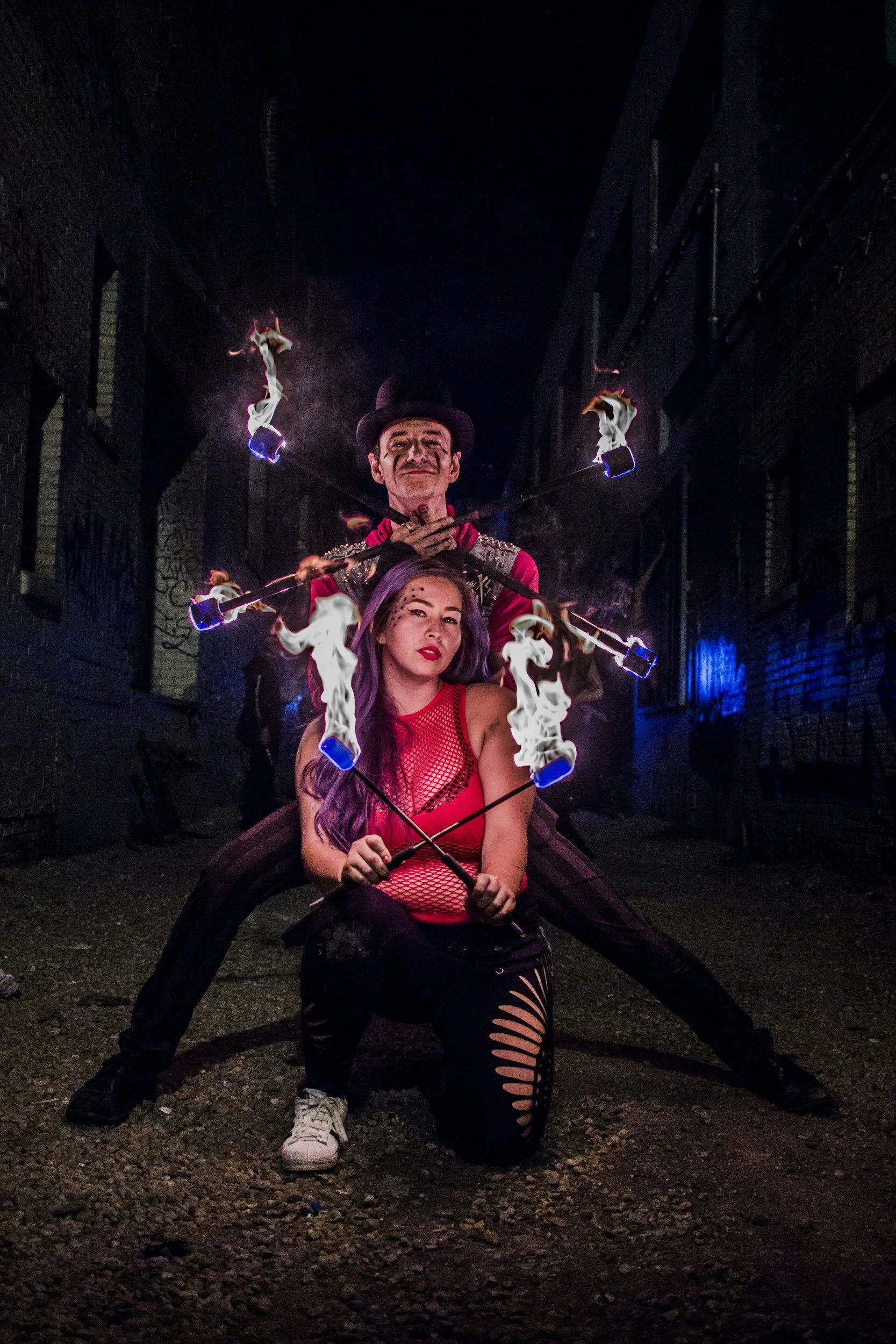 Two performers, a man and a woman, are in an alleyway at night. The woman is kneeling in front, holding lit fire poi sticks. The man is standing behind her, also holding fire poi sticks crossed in front of him. The alley is dark with graffiti on the walls.