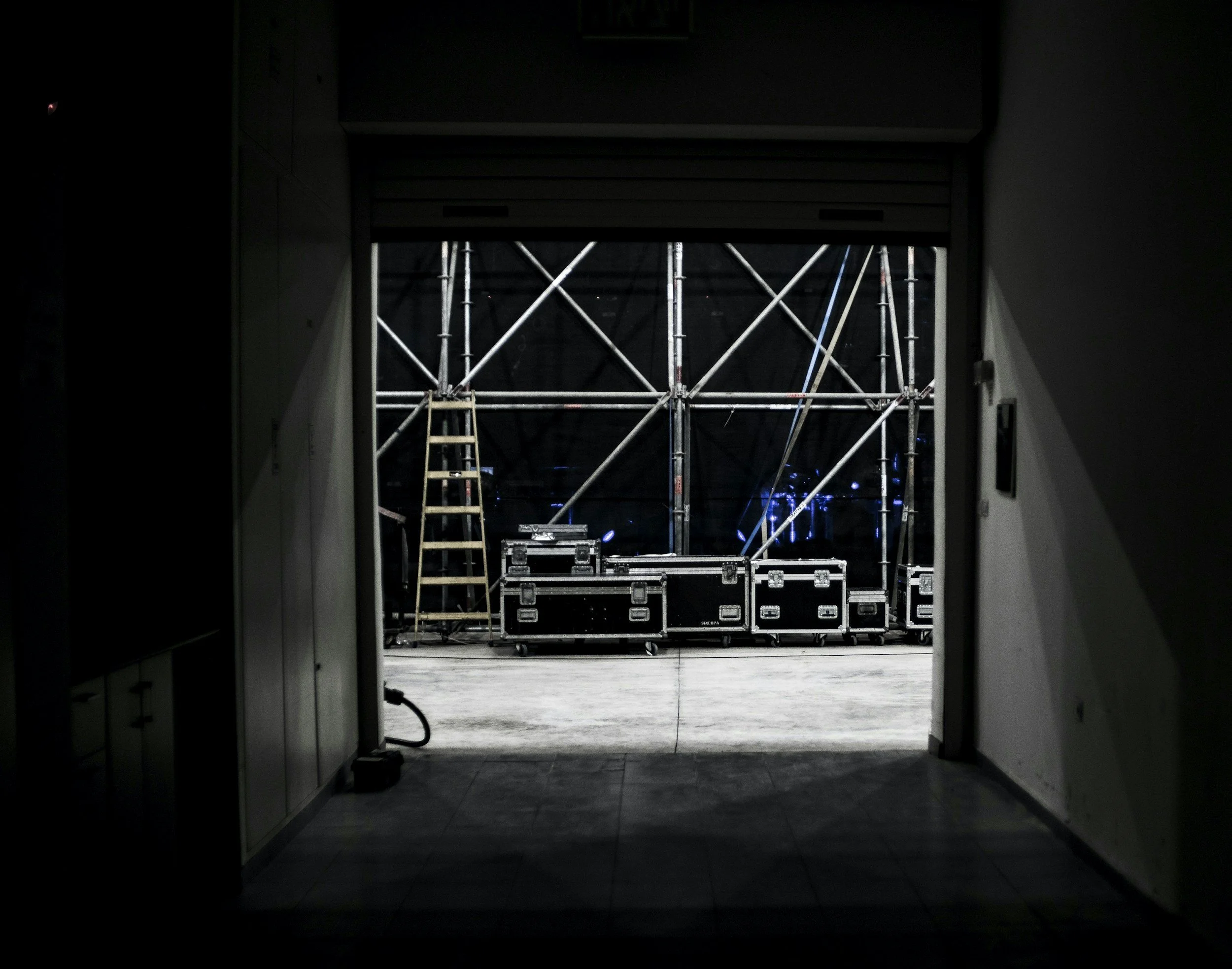 View of a backstage area at a concert or event, with scaffolding, a ladder, and equipment cases visible through an open doorway.