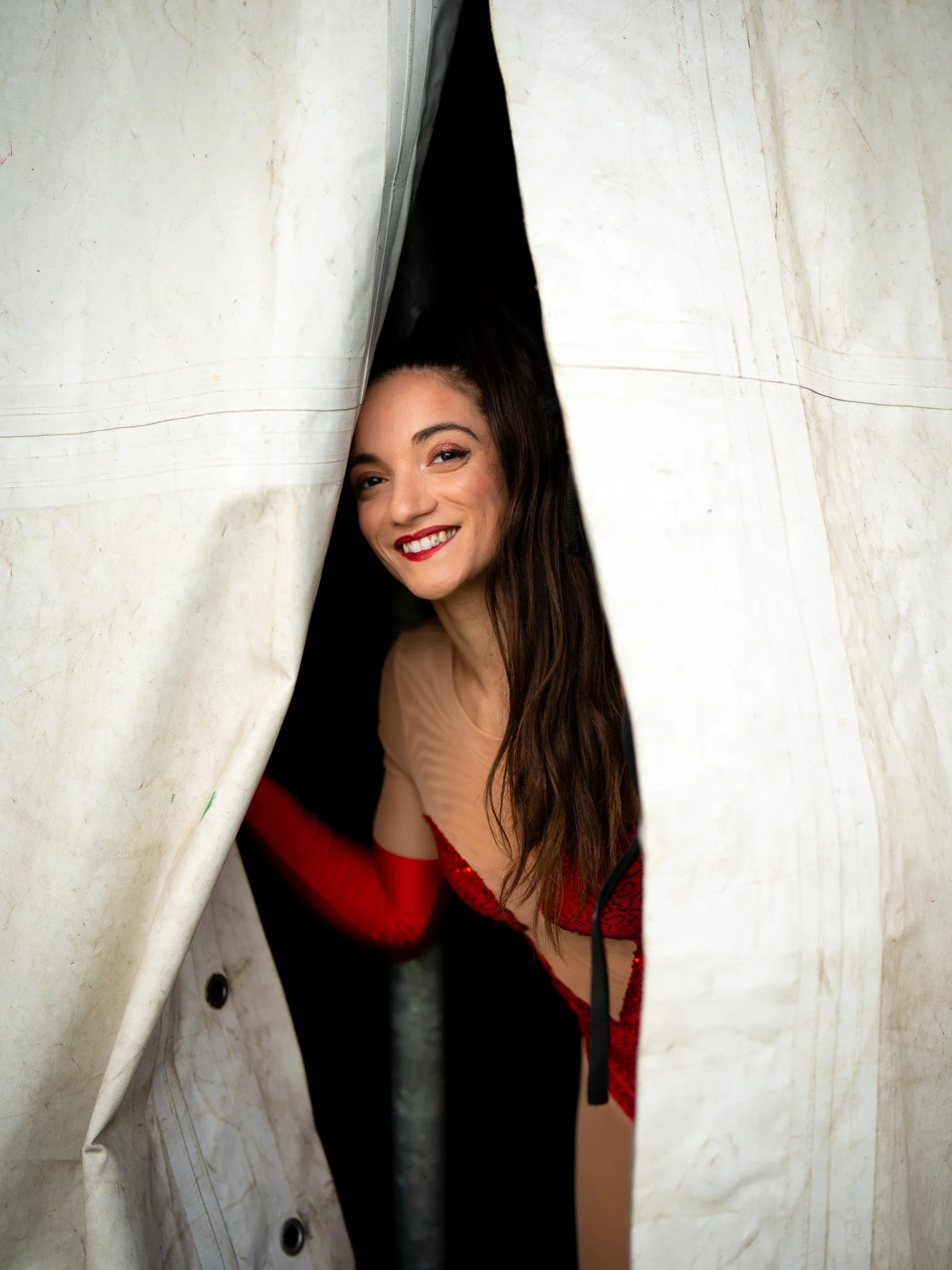 Woman with long dark hair and red lipstick, smiling and looking through a torn white curtain.