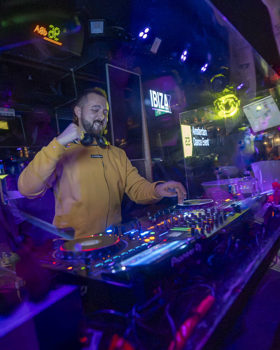 DJ performing at a nightclub with colorful LED lights, a sign that reads 'IBIZA', and a screen displaying 'Amsterdam Dance Event'.