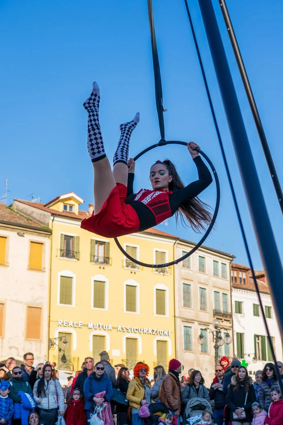A female performer is flying on a circular aerial hoop in front of a crowd during an outdoor event. She is wearing a red dress with a black and red top, black sleeves, and knee-high socks with a black and white checkered pattern. The crowd of onlookers is dressed warmly, with some wearing colorful costumes and accessories, in front of historic buildings with yellow facades and the sign "REALE MUTUA ASSICURAZIONI." The sky is clear and blue.