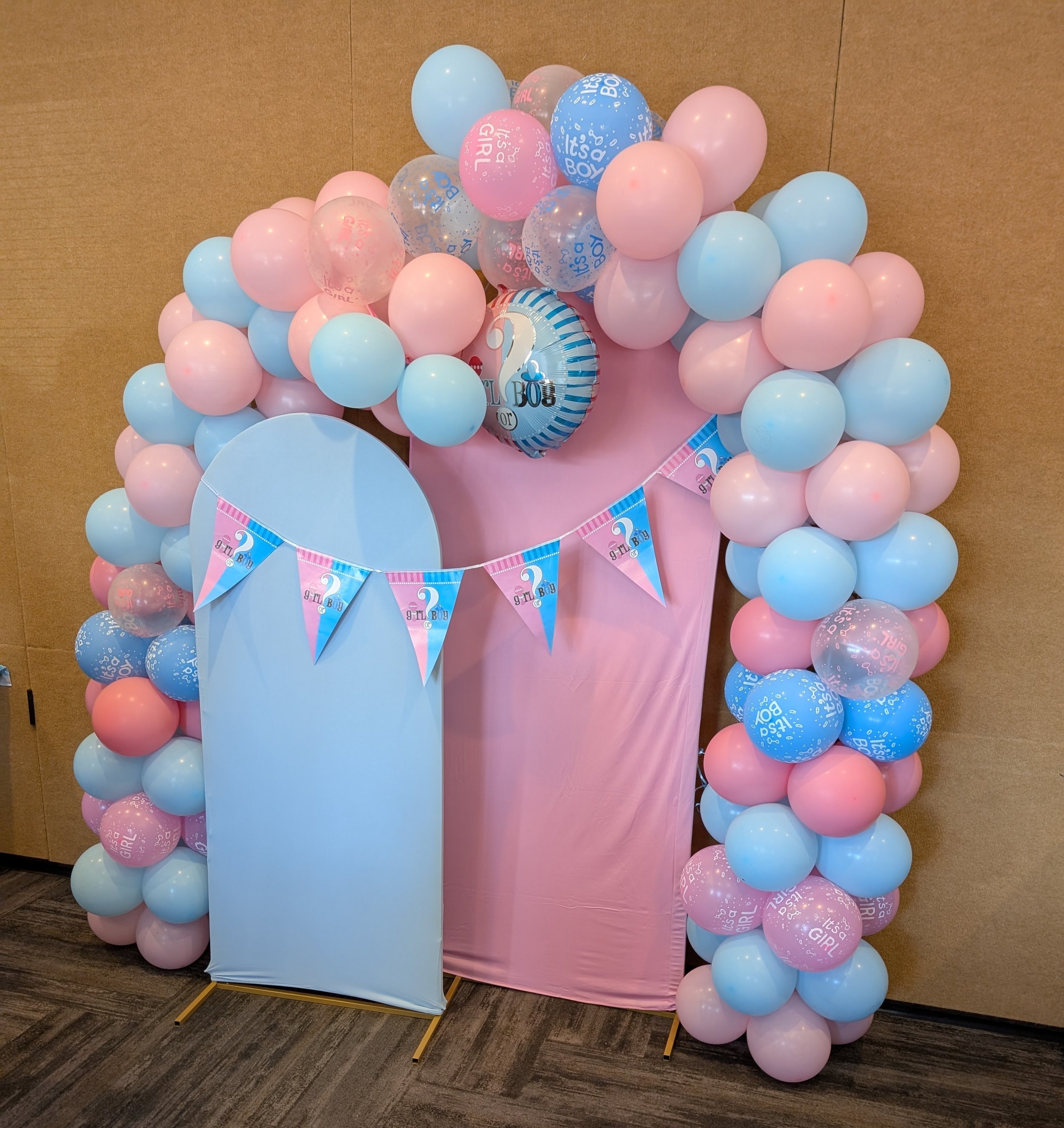 A balloon decoration for a gender reveal event with pink and blue balloons arranged around a pink and light blue backdrop featuring a ‘It's a Girl’ and ‘It's a Boy’ balloons banner for $125