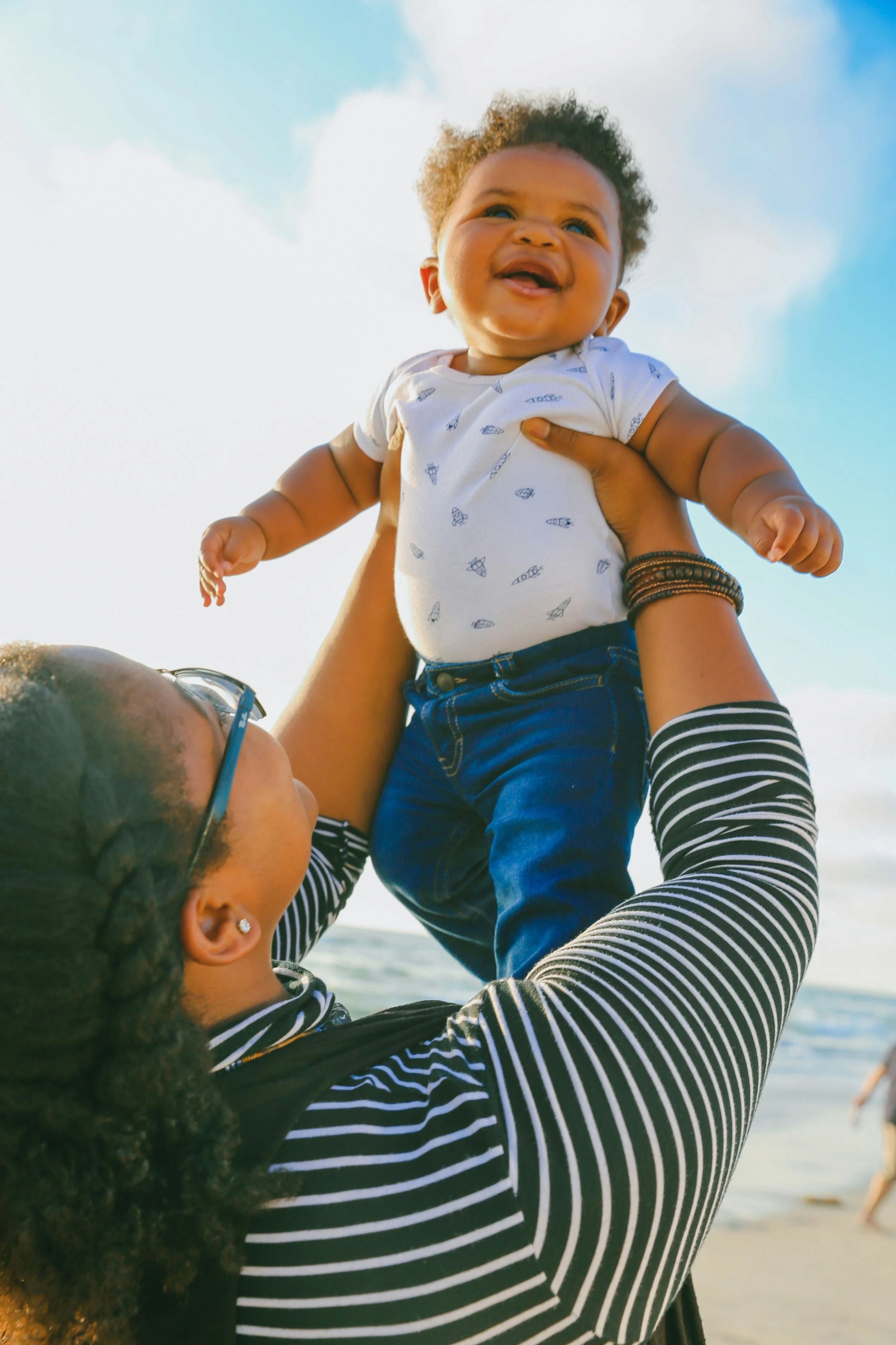 A woman with curly dark hair, wearing striped shirt and glasses, lifting a smiling toddler with brown skin, curly hair, and wearing a white T-shirt with small prints, at the beach with a clear blue sky in the background.