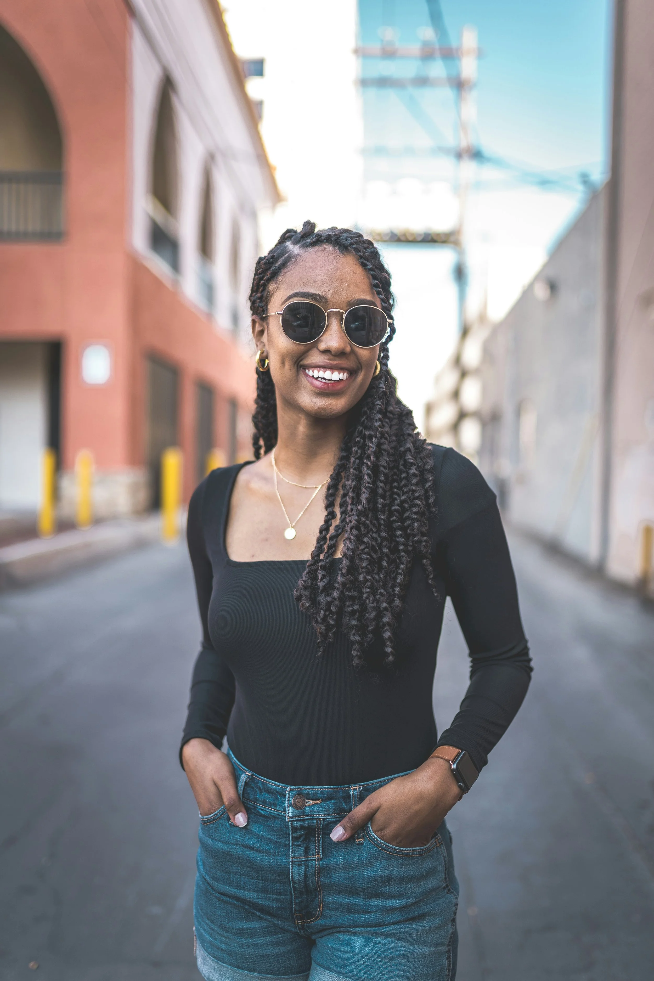 A young woman with styled braids, sunglasses, and gold jewelry smiling at the camera in an urban alleyway with colorful buildings and utility wires.