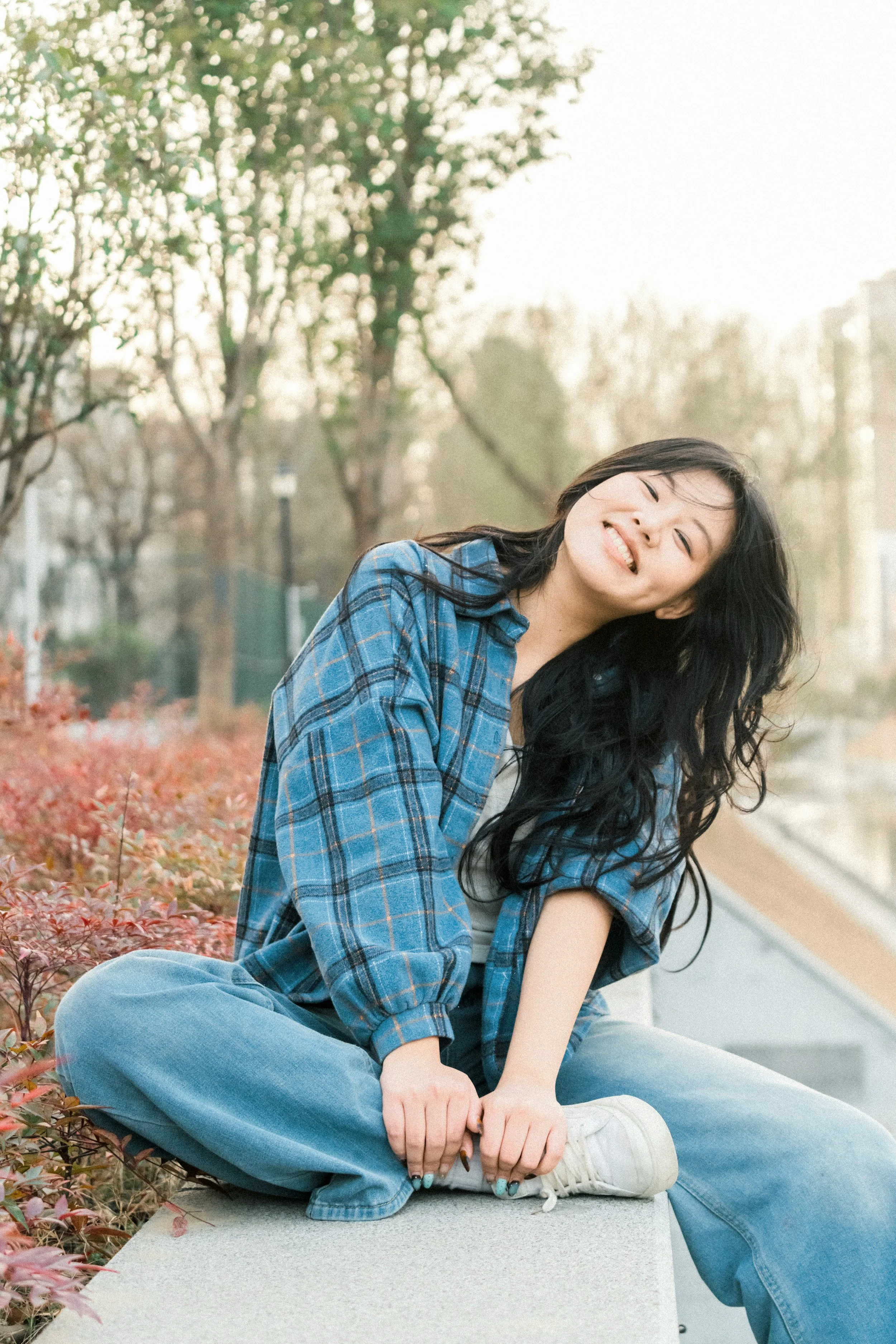 Young woman with long dark hair sitting on a ledge outdoors, smiling, wearing a blue plaid shirt and light blue jeans, surrounded by autumn leaves, in a park with trees in the background.