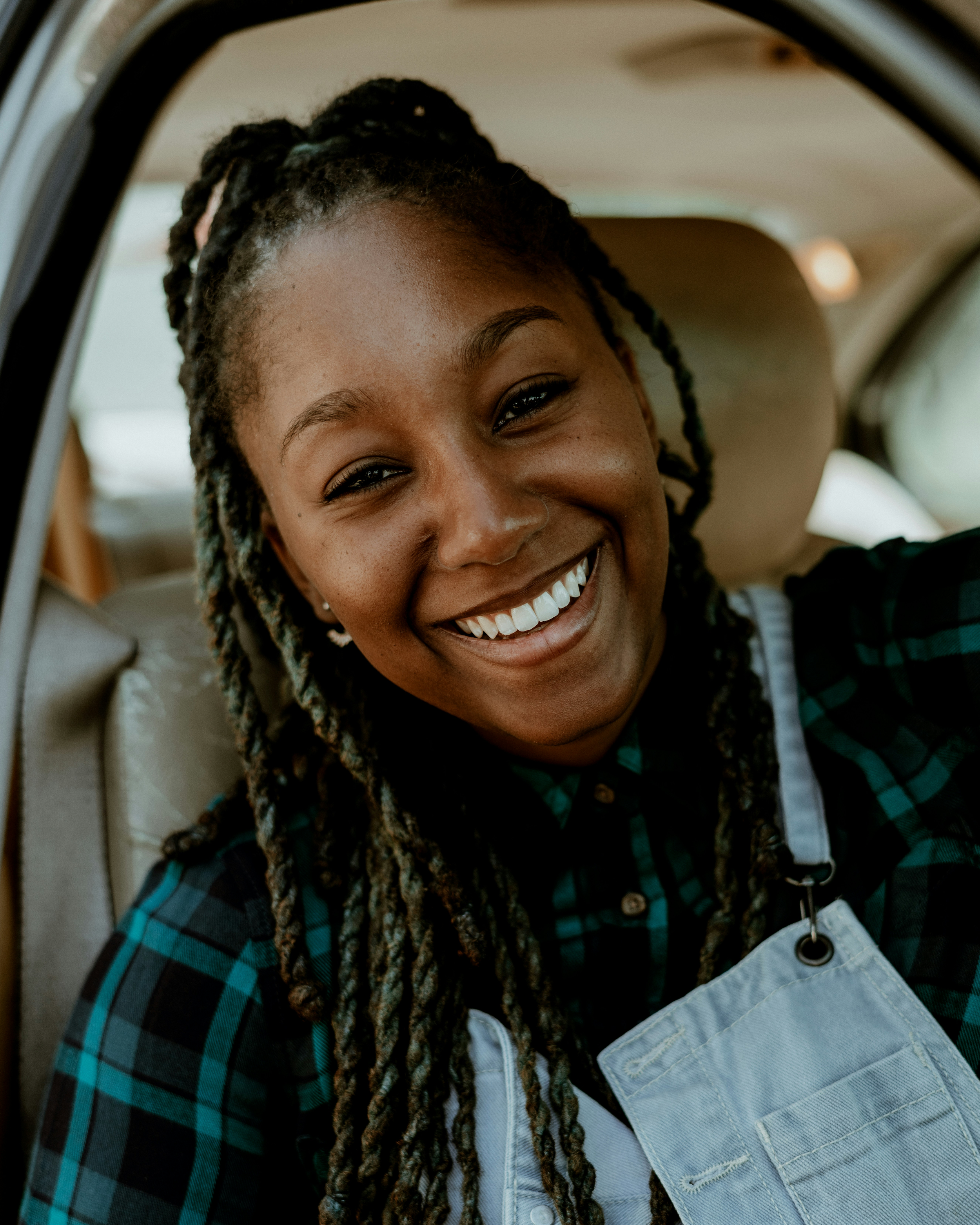 Close-up of a smiling woman with long dreadlocks wearing a plaid shirt and a white apron, sitting inside a vehicle.