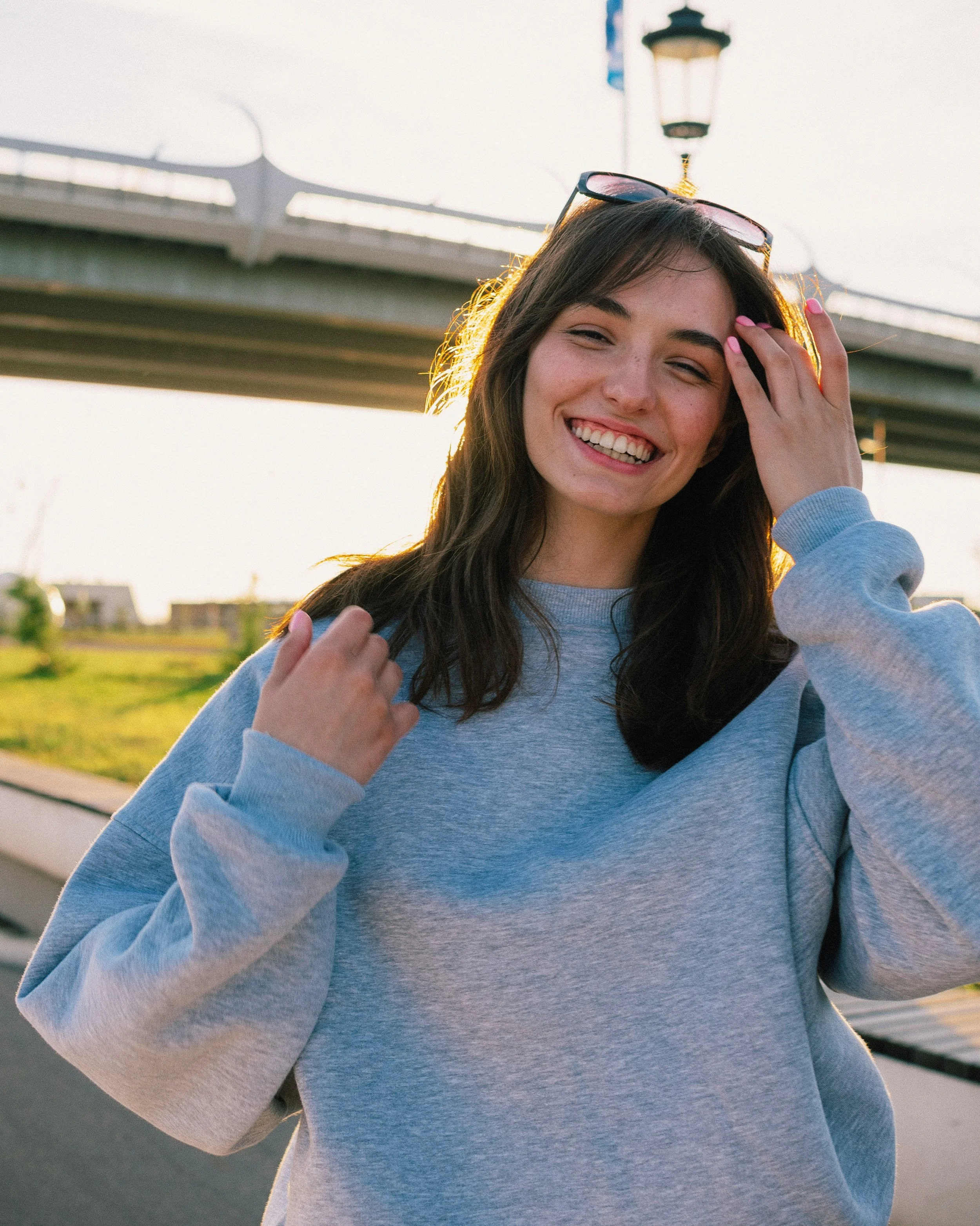 A young woman smiling outdoors during daytime, wearing a light gray sweatshirt, with sunglasses resting on her head, standing near a bridge and lamp post in the background.