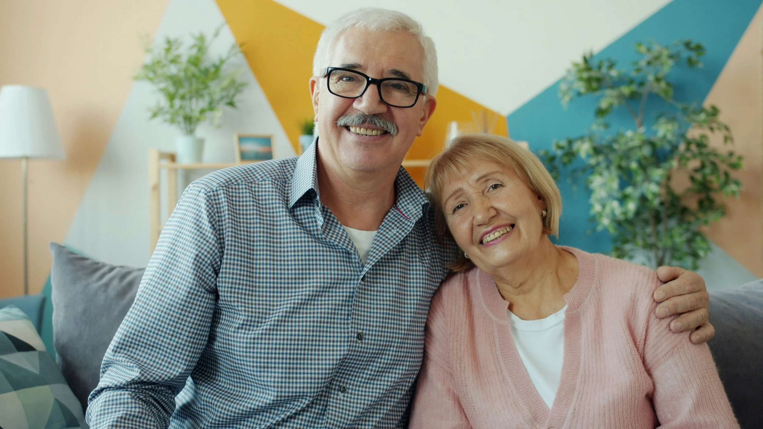 An elderly couple sitting together on a couch in a brightly decorated living room, smiling at the camera. The man has white hair, glasses, and a checkered shirt, while the woman has light brown hair and is wearing a pink sweater.