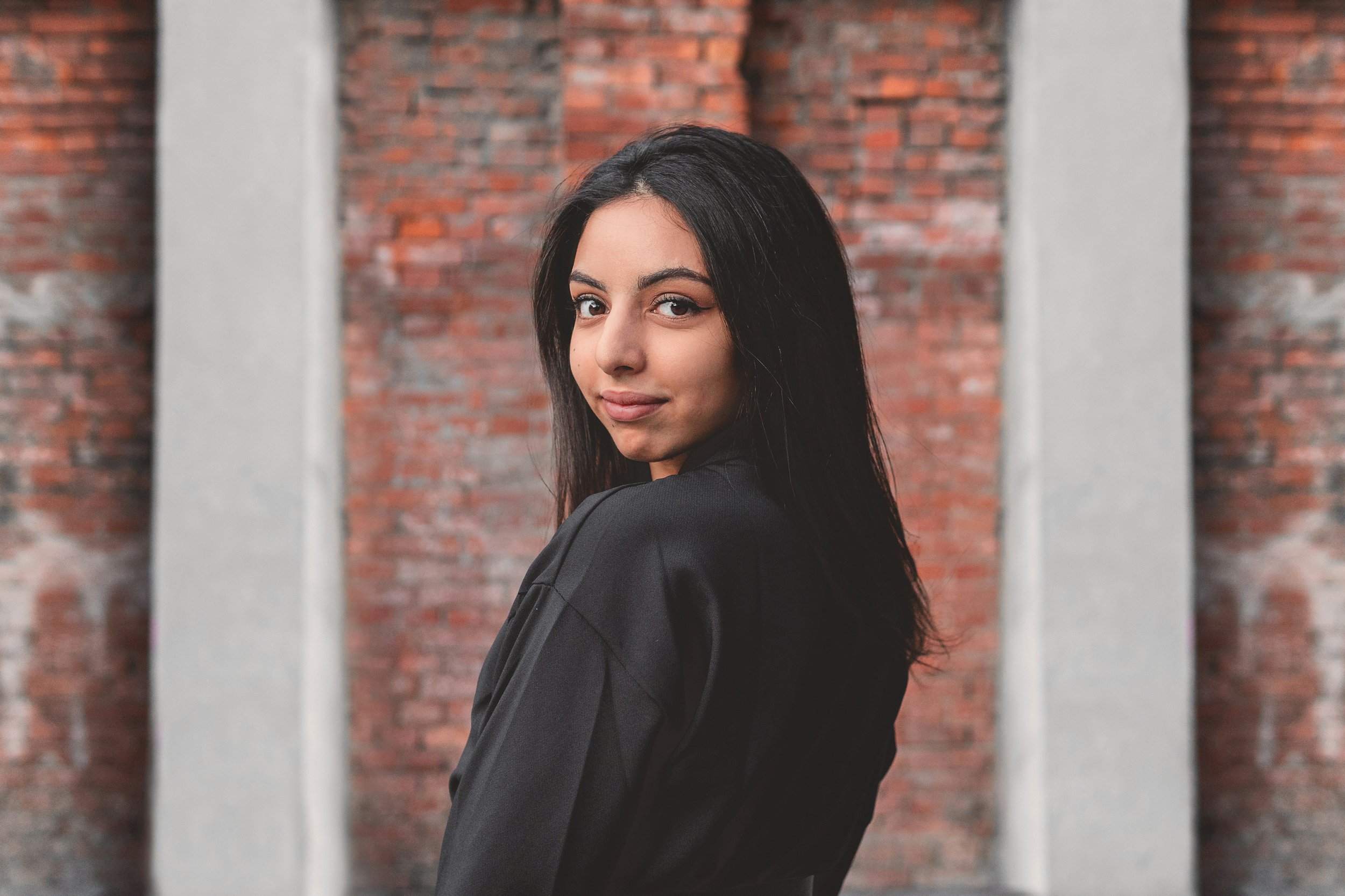 A woman with long dark hair and light skin standing against a red brick wall with white columns, looking over her shoulder with a slight smile.