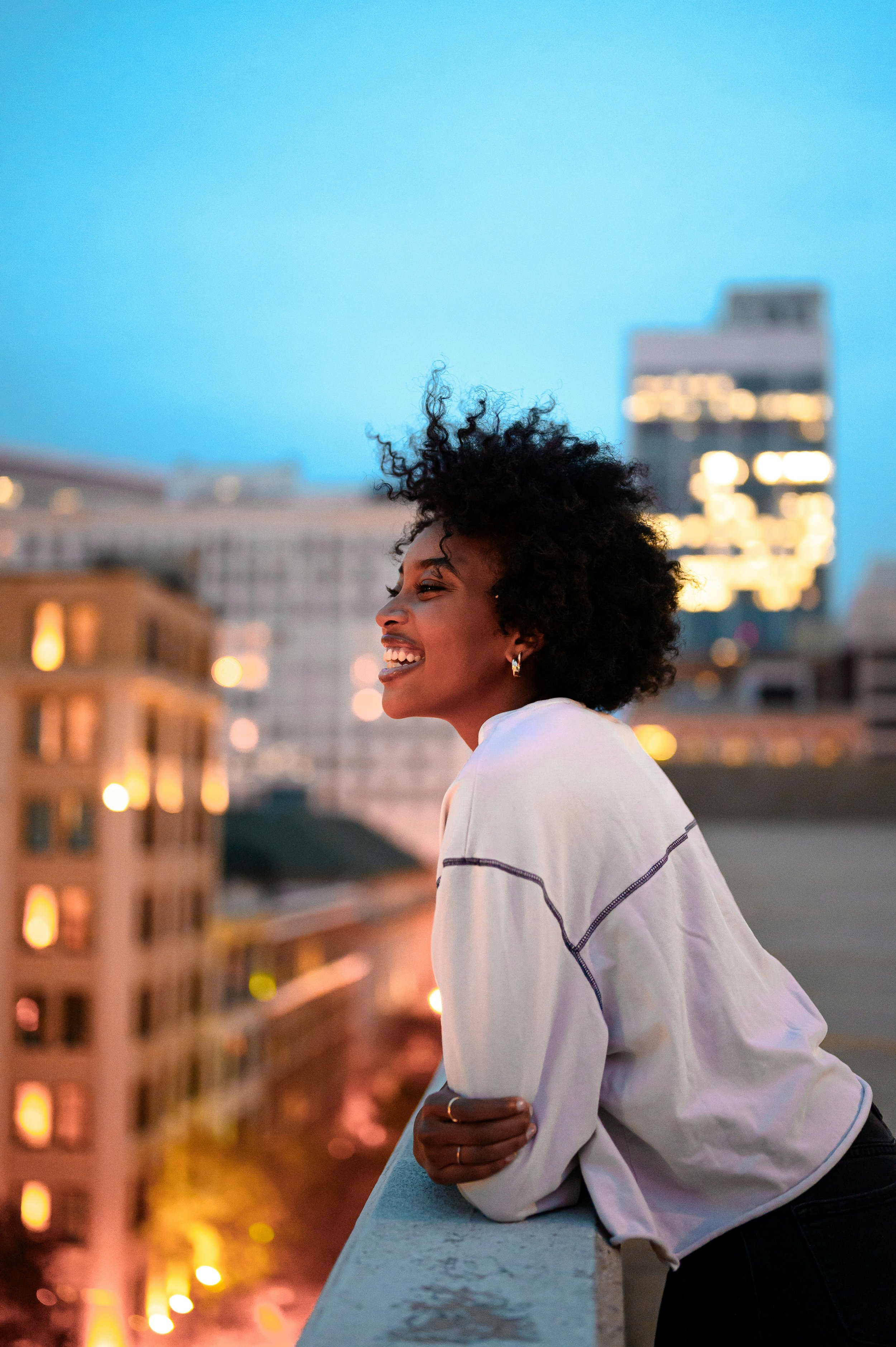 A smiling woman with curly black hair wearing a white sweatshirt with black trim, leaning on a concrete railing on a city rooftop at dusk, with a blurred cityscape and tall buildings with lit windows in the background.