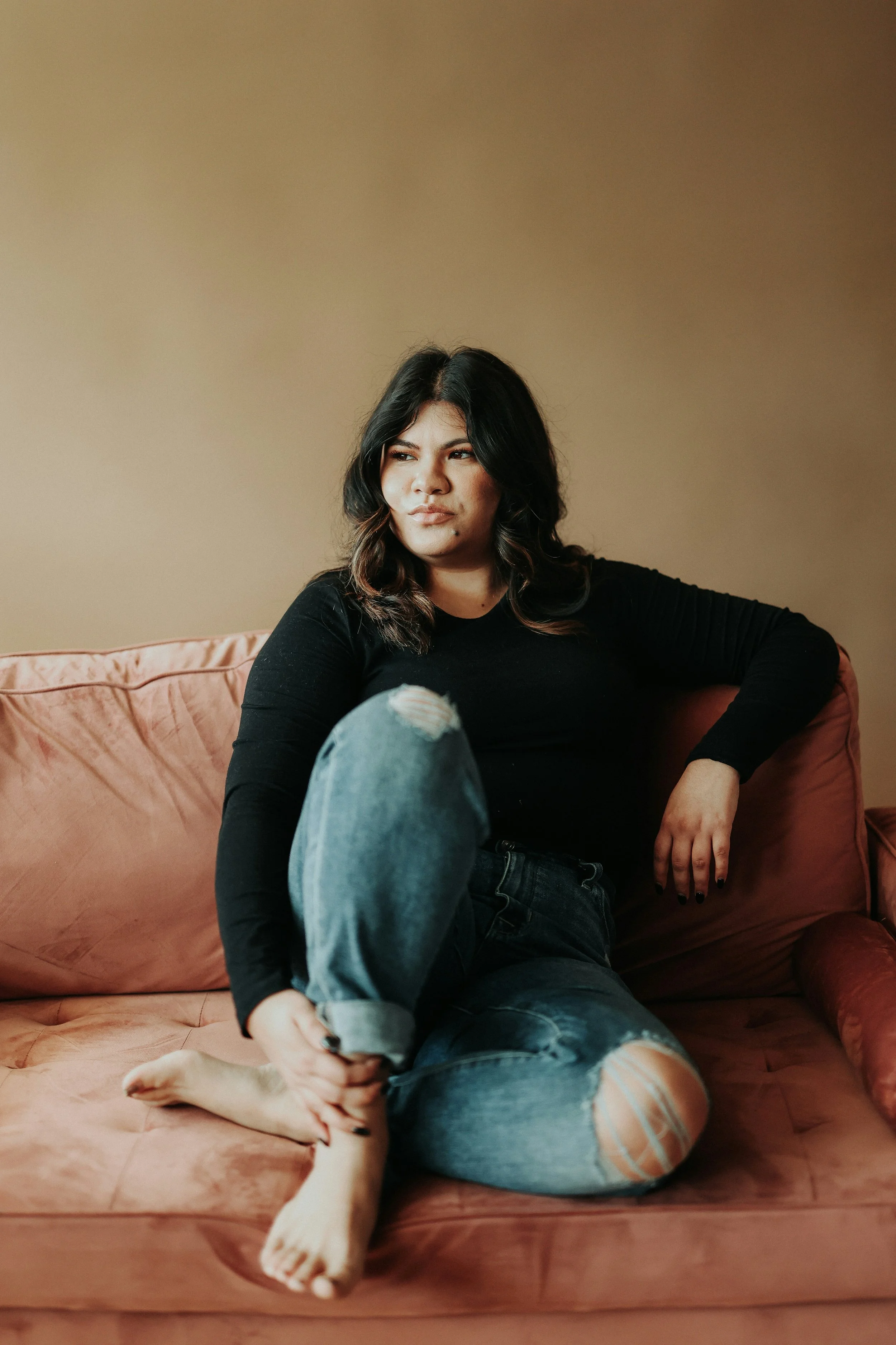 A woman sitting on a pink couch, wearing a black long sleeve top and ripped jeans, with dark, wavy hair, looking to the side with a serious expression.
