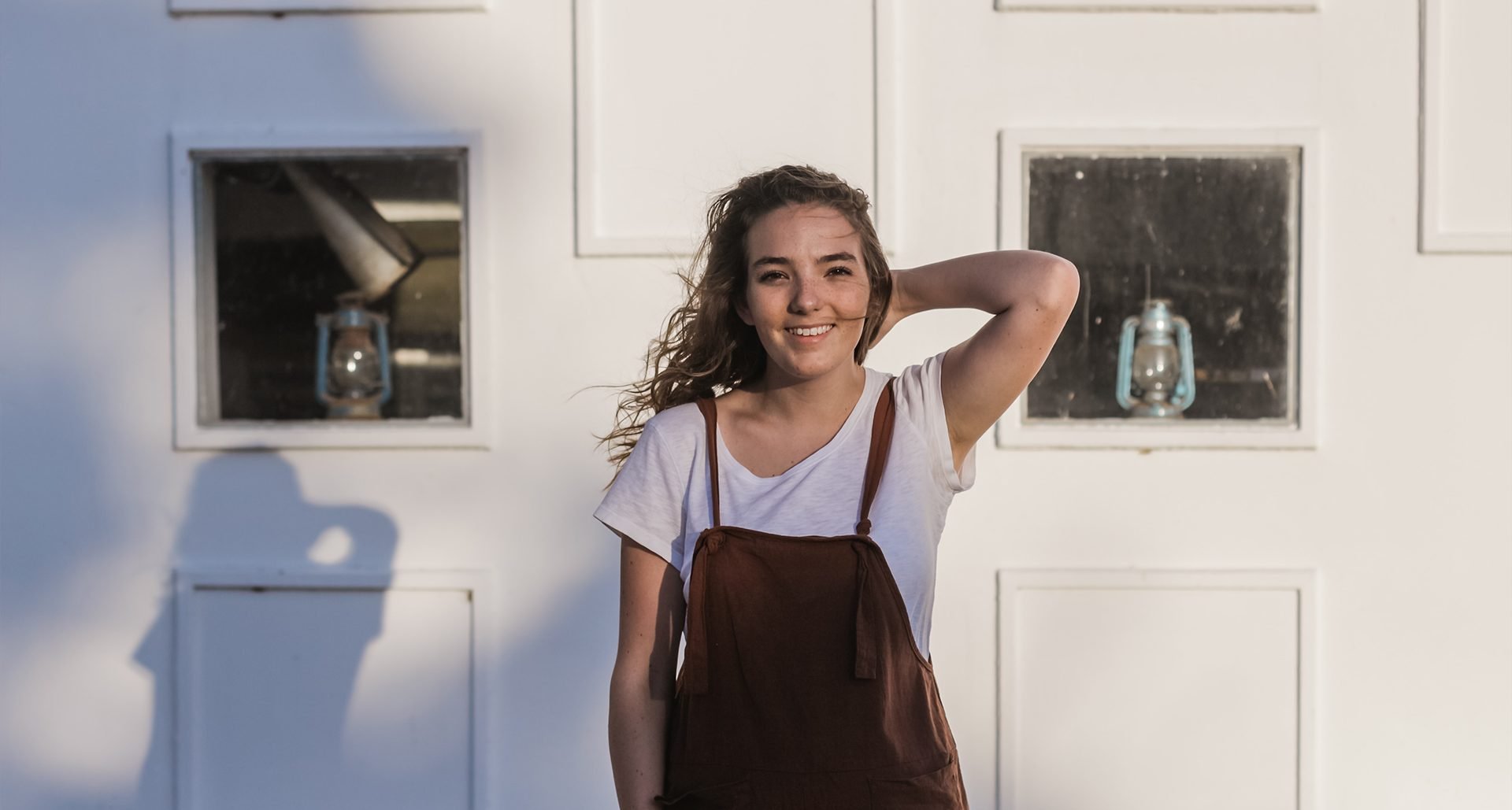 A young woman with curly brown hair smiling, standing in front of a white wall with two rectangular windows that have black screens and lanterns inside.