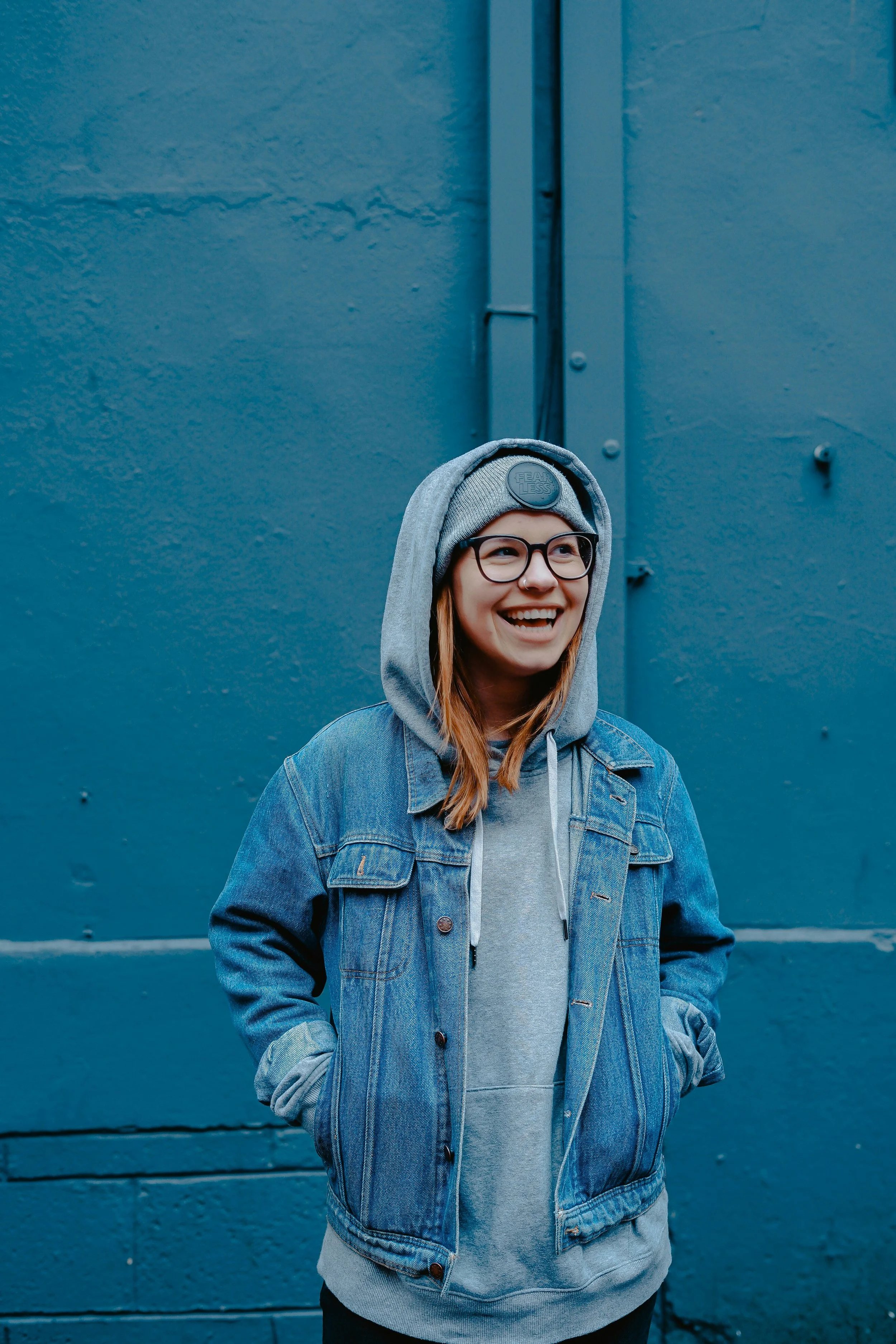 A young woman with glasses and a nose piercing smiling, wearing a gray hoodie and denim jacket, standing against a blue wall.