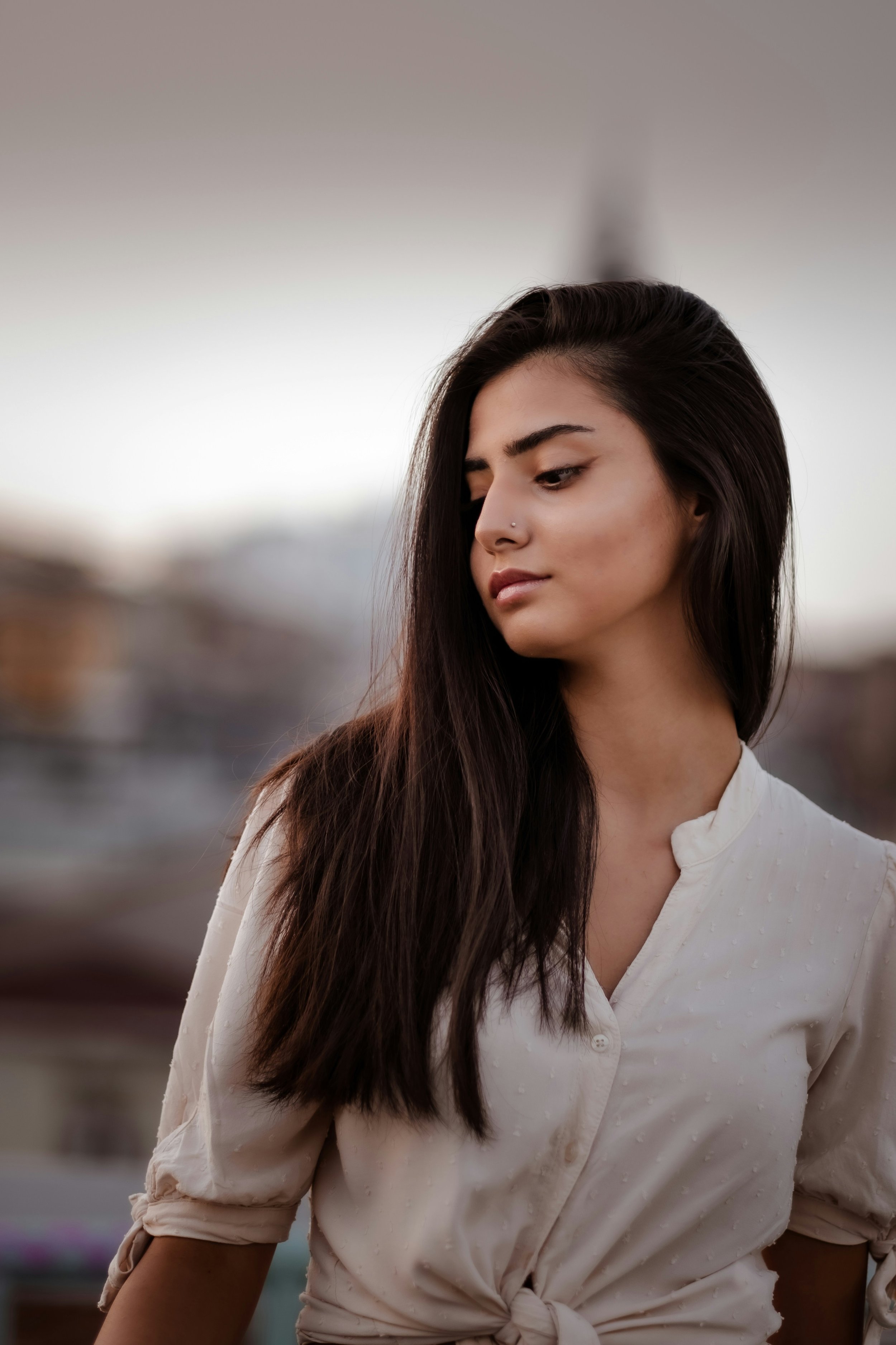 A woman with long dark hair and a nose piercing wearing a white button-up shirt with rolled-up sleeves, standing outdoors during sunset.