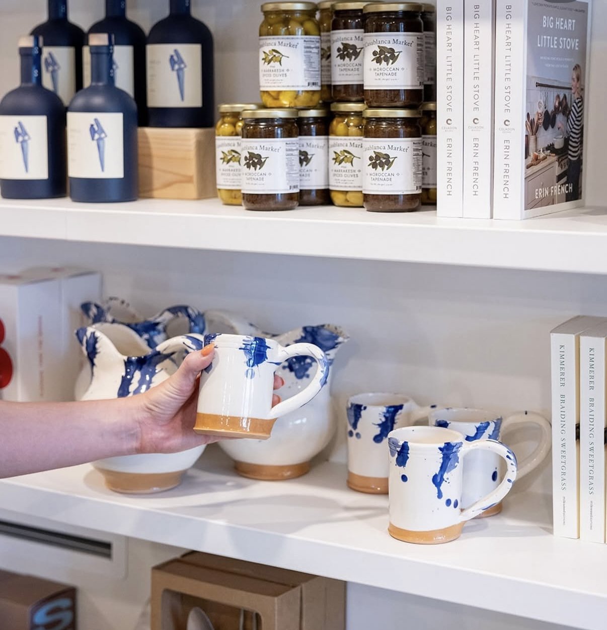 A white shelf displaying jars of olives and preserved vegetables, with a row of stacked books on the right, and hand holding a ceramic mug with blue paint splashes and a tan bottom, surrounded by matching mugs and bowls also with blue splashes, on a