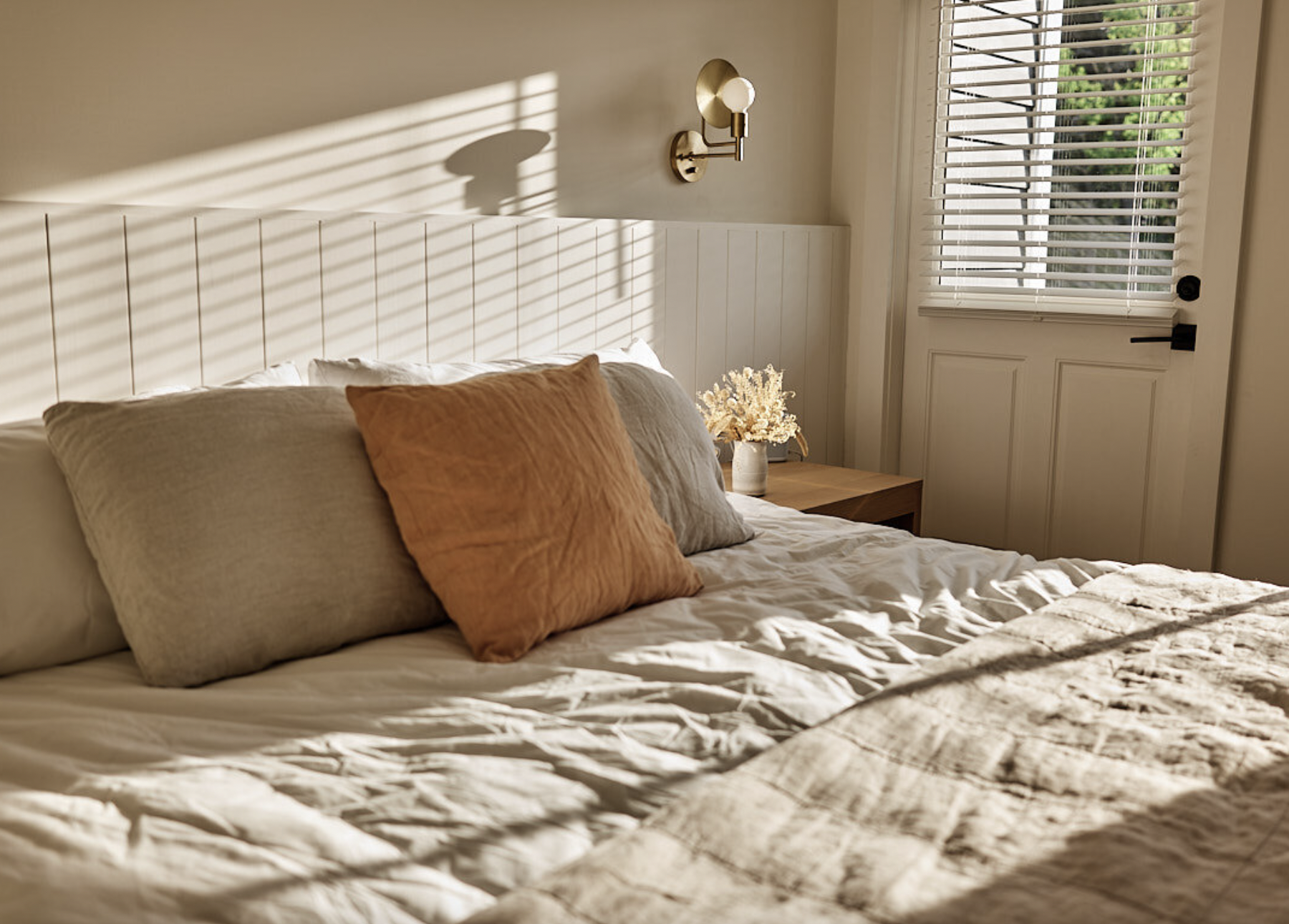 A neatly made bed with beige and rusty colored pillows, sunlight streaming through window blinds casting shadows on the bed and wall, with a wall-mounted light fixture and a small side table with a decorative dried flower arrangement near the window.