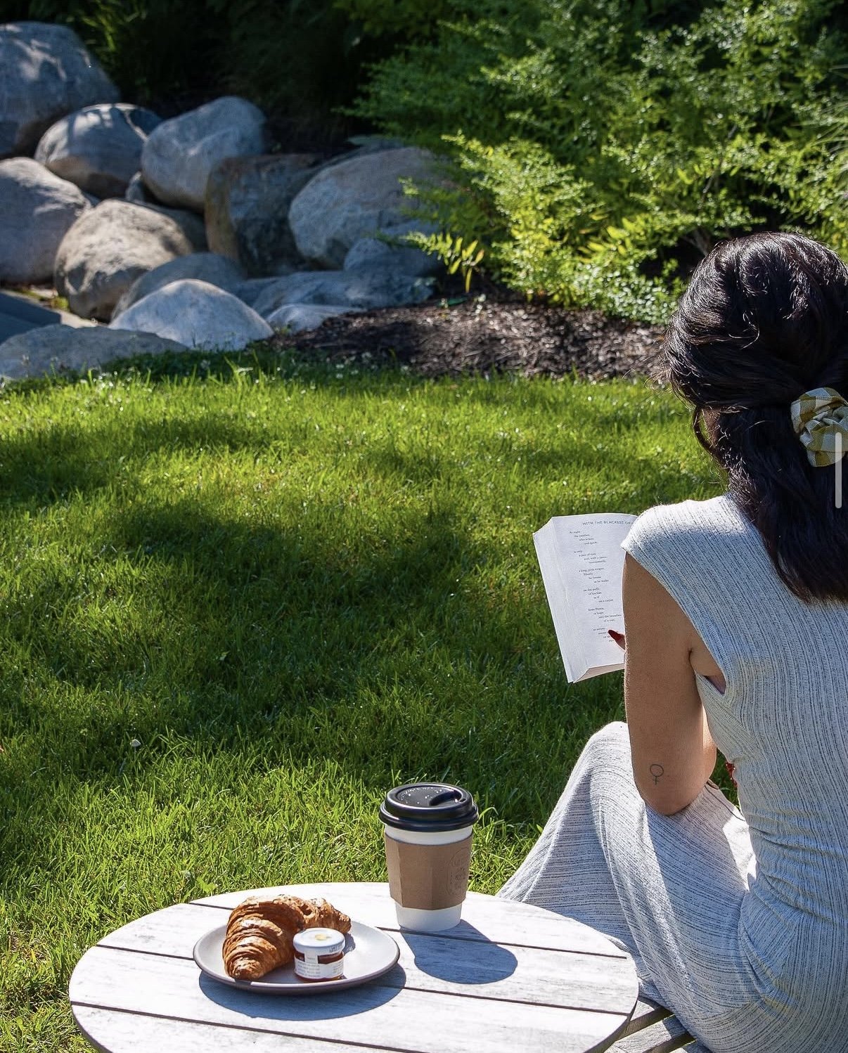 A woman sitting outdoors on grass, reading a book, with a small round table in front of her holding a coffee cup and a plate with a croissant and jam, surrounded by greenery and rocks.