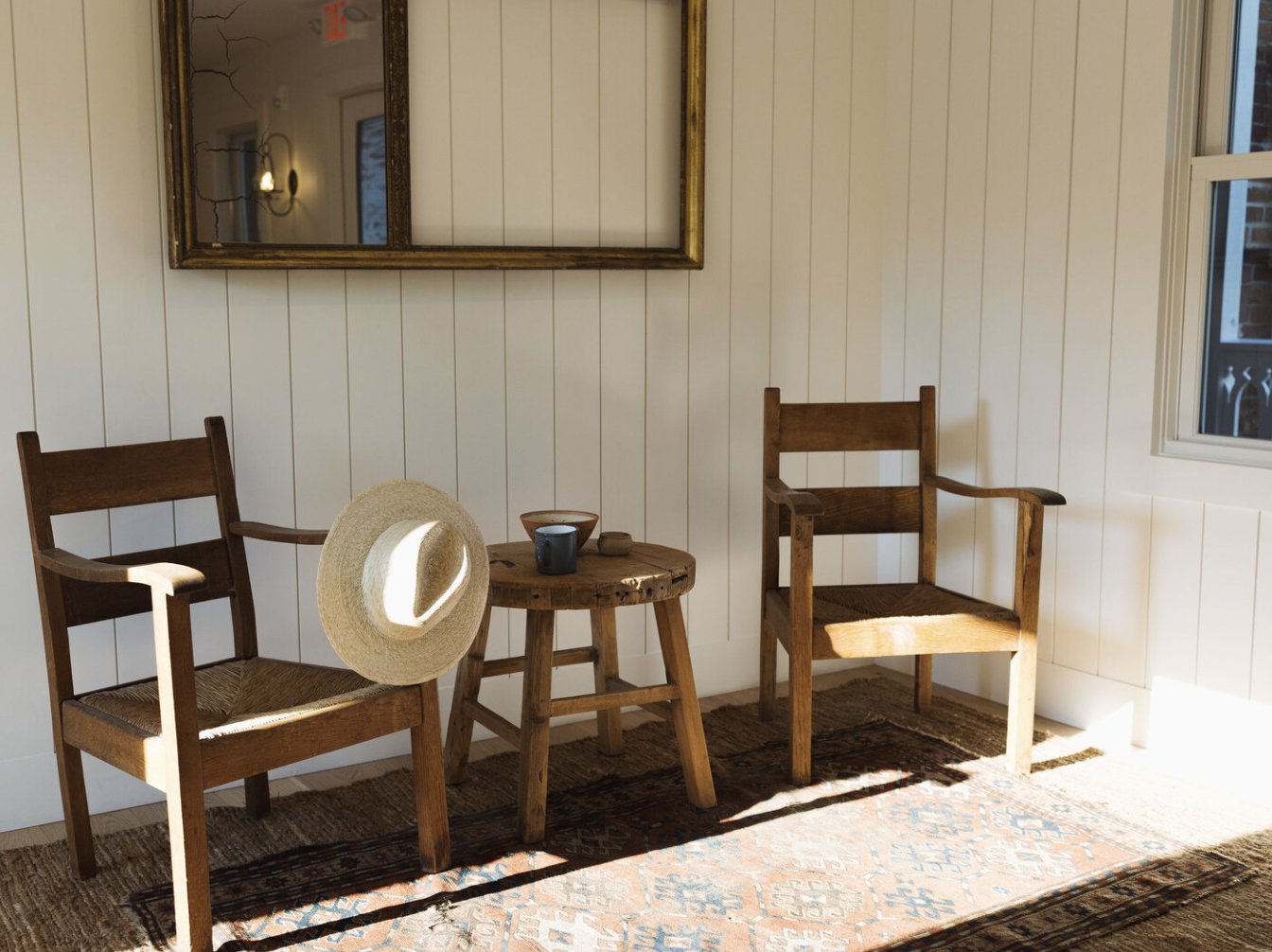 Sunlit corner with two wooden chairs, a small round wooden table, a straw hat hanging on one chair, a blue cup, and a rug on the floor, with a mirror and window on the white paneled wall.