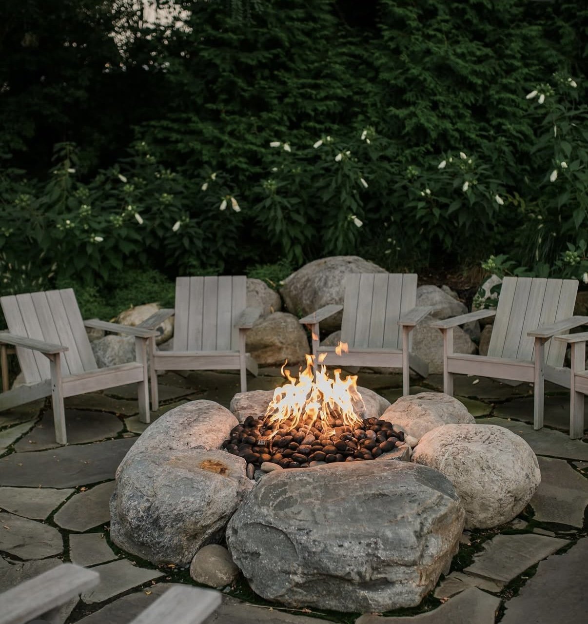 Outdoor fire pit surrounded by four wooden chairs in a garden setting with large rocks and dense green foliage in the background.