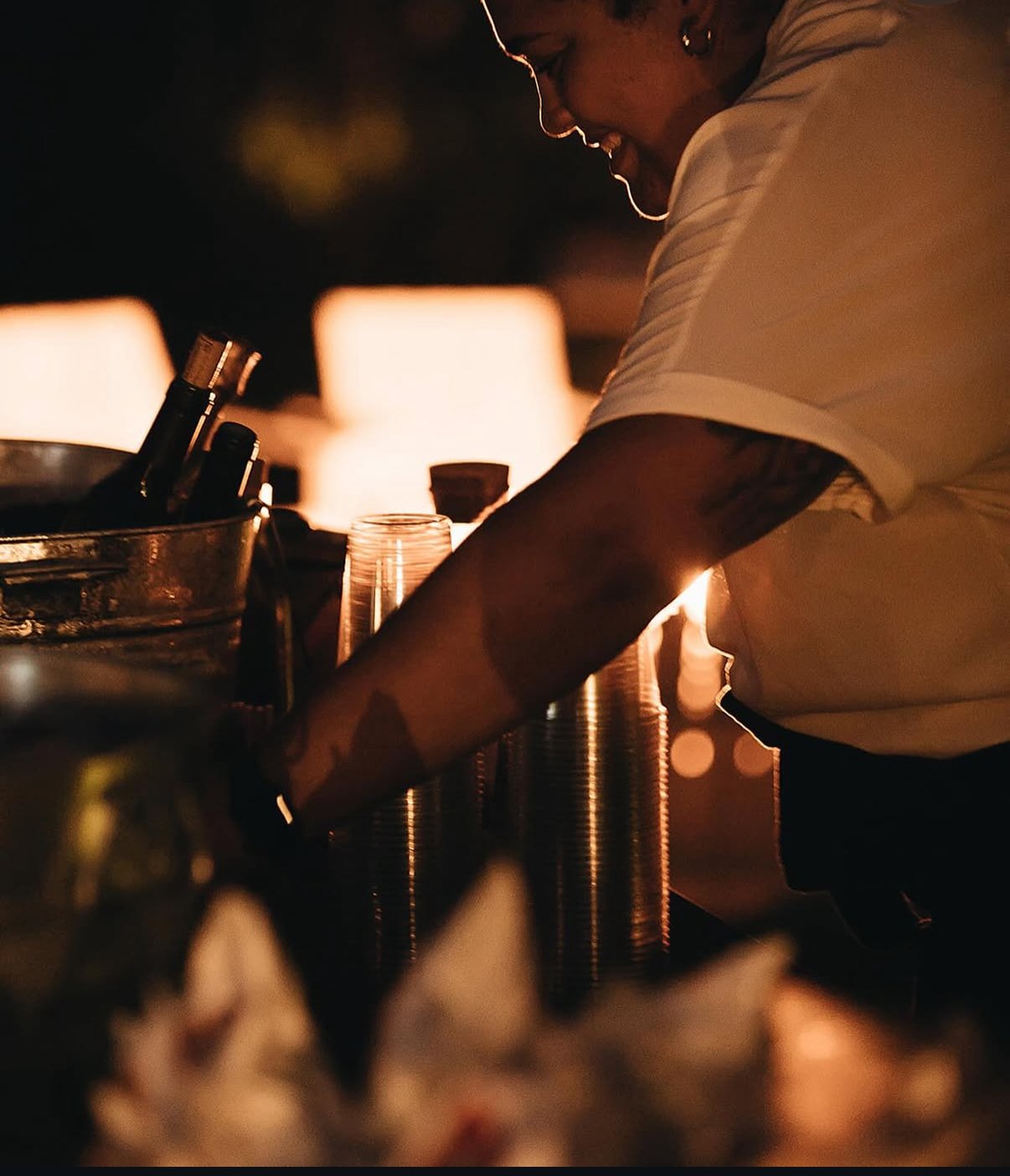 A person preparing drinks at a bar during nighttime, with warm lighting and glasses on the counter.