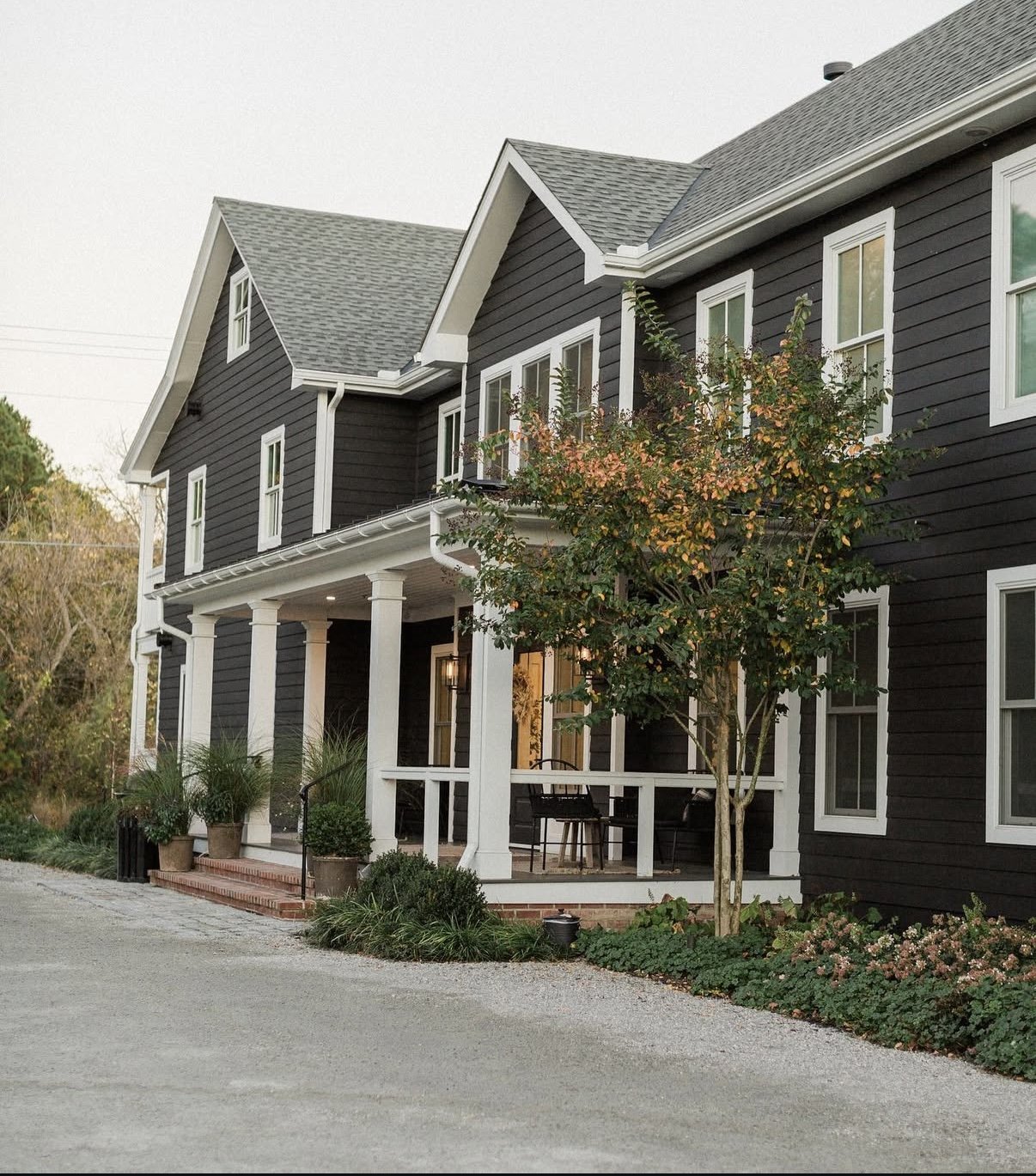 A two-story black house with white trim and a front porch, surrounded by greenery, and with a gravel driveway.
